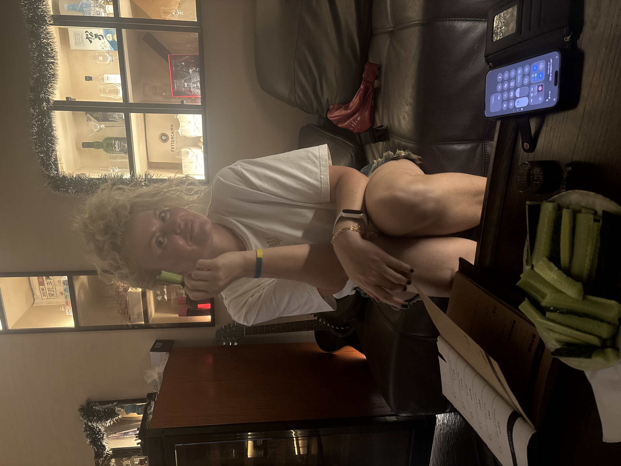 A woman with curly blonde hair and a white t-shirt sits on a dark leather couch in a dimly lit room. She is holding a small green object to her cheek and looking towards the camera. She is wearing a blue and yellow wristband. In front of her is a dark wooden table with a menu, a plate of sliced cucumbers, and a mobile phone displaying its control center interface. Behind her, a guitar leans against a dark wooden cabinet. The background features a lit display cabinet filled with bottles and glasses, decorated with festive tinsel. The scene appears to be a bar or lounge in Nanhai District, China.