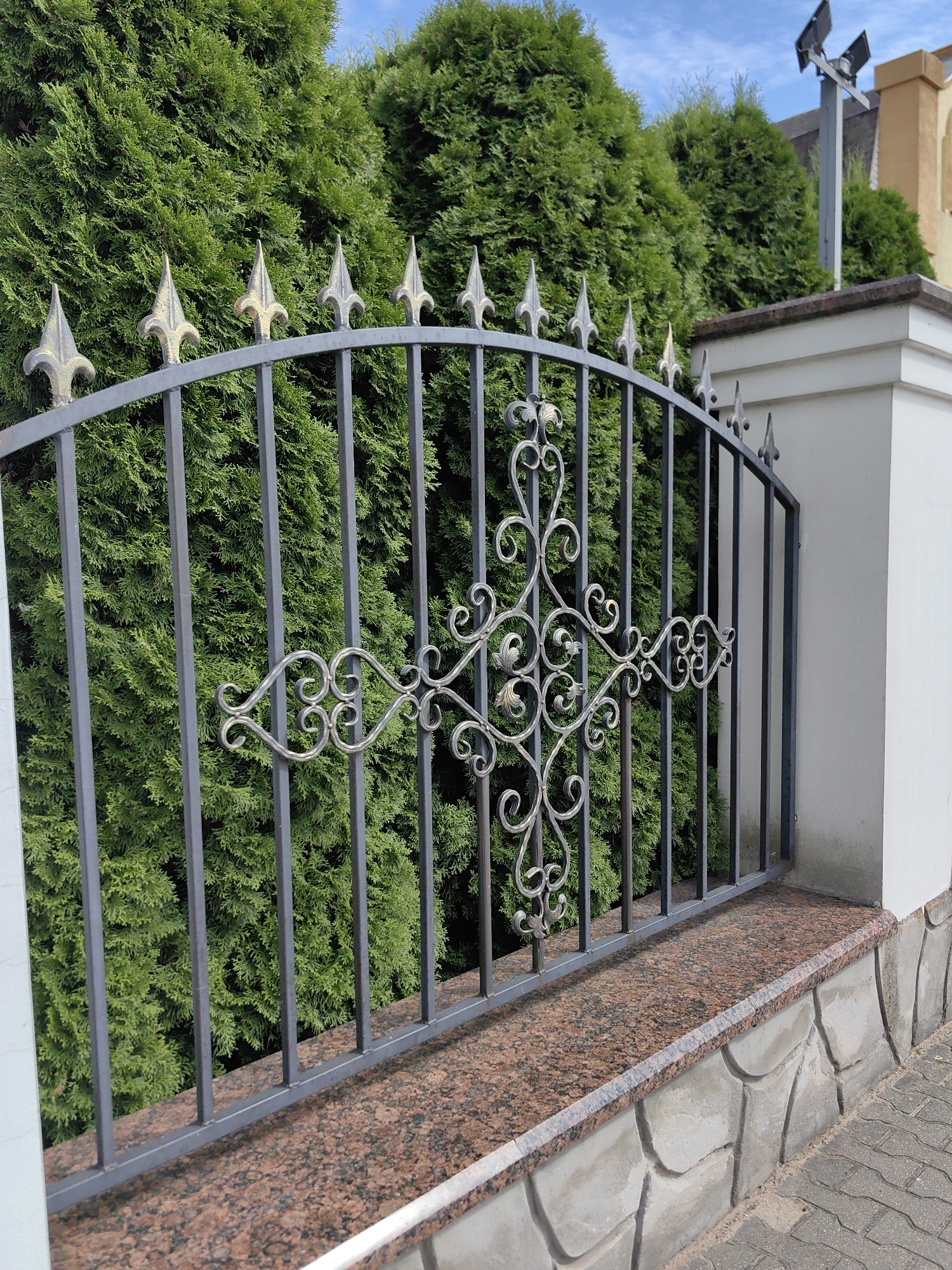 An ornate dark grey metal fence with decorative scrollwork and pointed finials stands in front of a dense green evergreen hedge. The fence is supported by white concrete pillars. A polished reddish-brown granite cap runs along the top of a stone-clad base. Paved bricks are visible on the ground in front of the fence. In the background, several solar-powered security lights are mounted on a pole above the hedge. The sky is blue with scattered white clouds. This scene appears to be a residential property in Pastavy, Belarus. There are no people visible in the image.