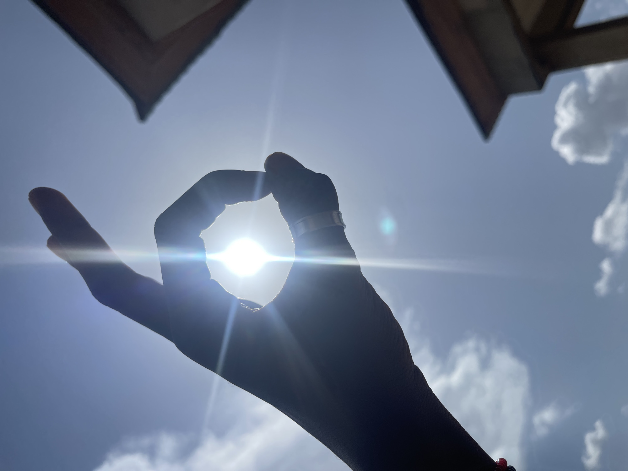A silhouette of a person's hand frames the bright sun against a clear blue sky. The sun is so intense that it creates lens flare and rays of light radiating outwards. The fingers of the hand are curled, with the thumb and index finger forming a circle around the sun. A ring is visible on one of the fingers, and a bracelet with red and black beads adorns the wrist.
In the upper corners of the frame, the eaves of a wooden structure are visible, suggesting an outdoor setting with some form of shelter. Puffy white clouds are scattered across the sky, indicating fair weather. The lighting suggests it is daytime, likely midday given the directness of the sun. The overall mood conveyed is one of warmth, light, and perhaps a sense of peace or mindfulness, as the hand gesture can be interpreted as an "OK" sign. The location context of Jos, Nigeria, provides a geographical anchor for the scene. There is no visible text in the image.