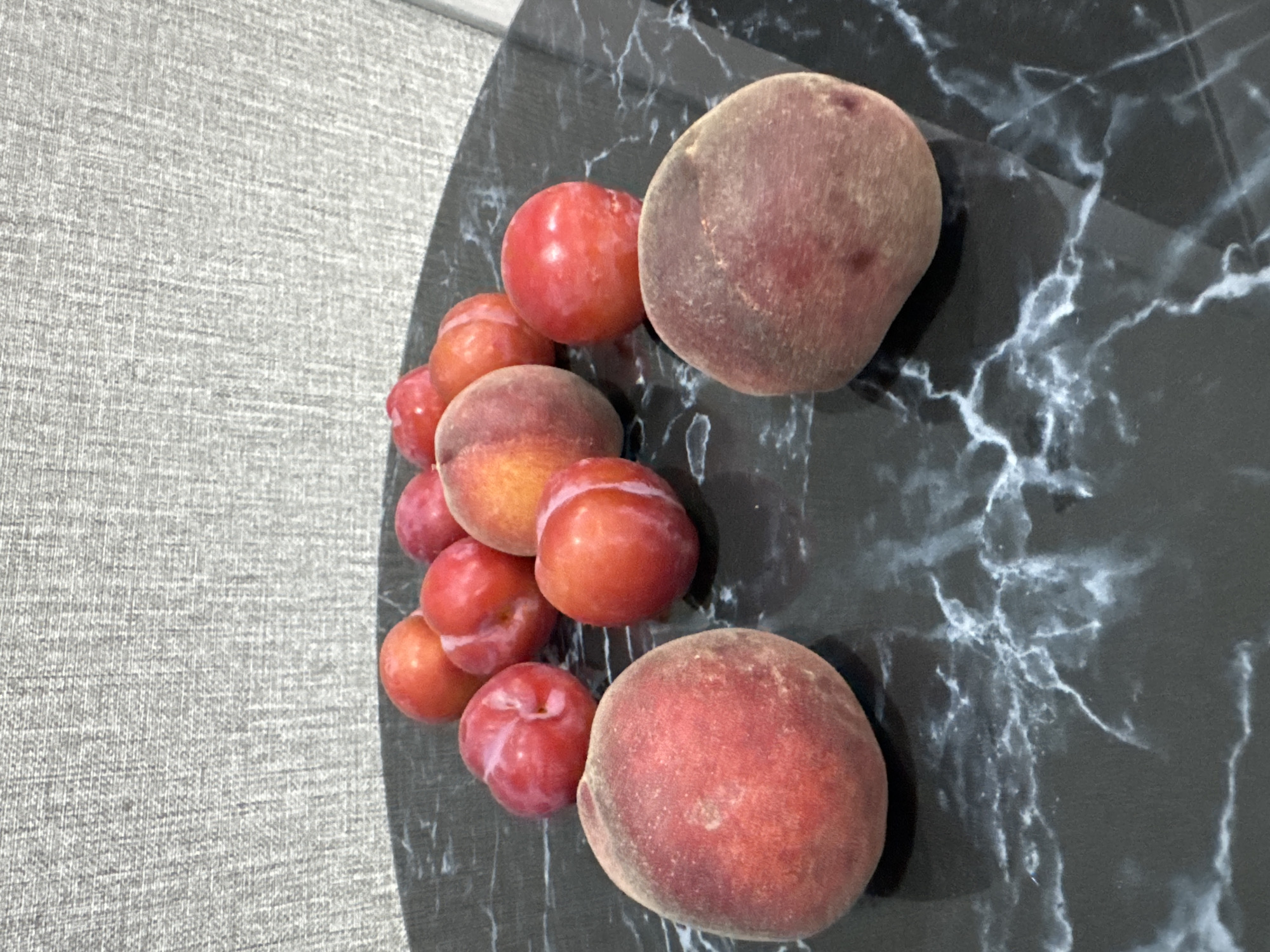 This image shows a collection of plums and two peaches arranged on a dark marble surface. The background is a textured grey fabric. The fruits are mostly red and pink, with some exhibiting a slight blush of yellow or orange. The lighting suggests an indoor setting. The location of Солонешное, Russia is not depicted in the image.