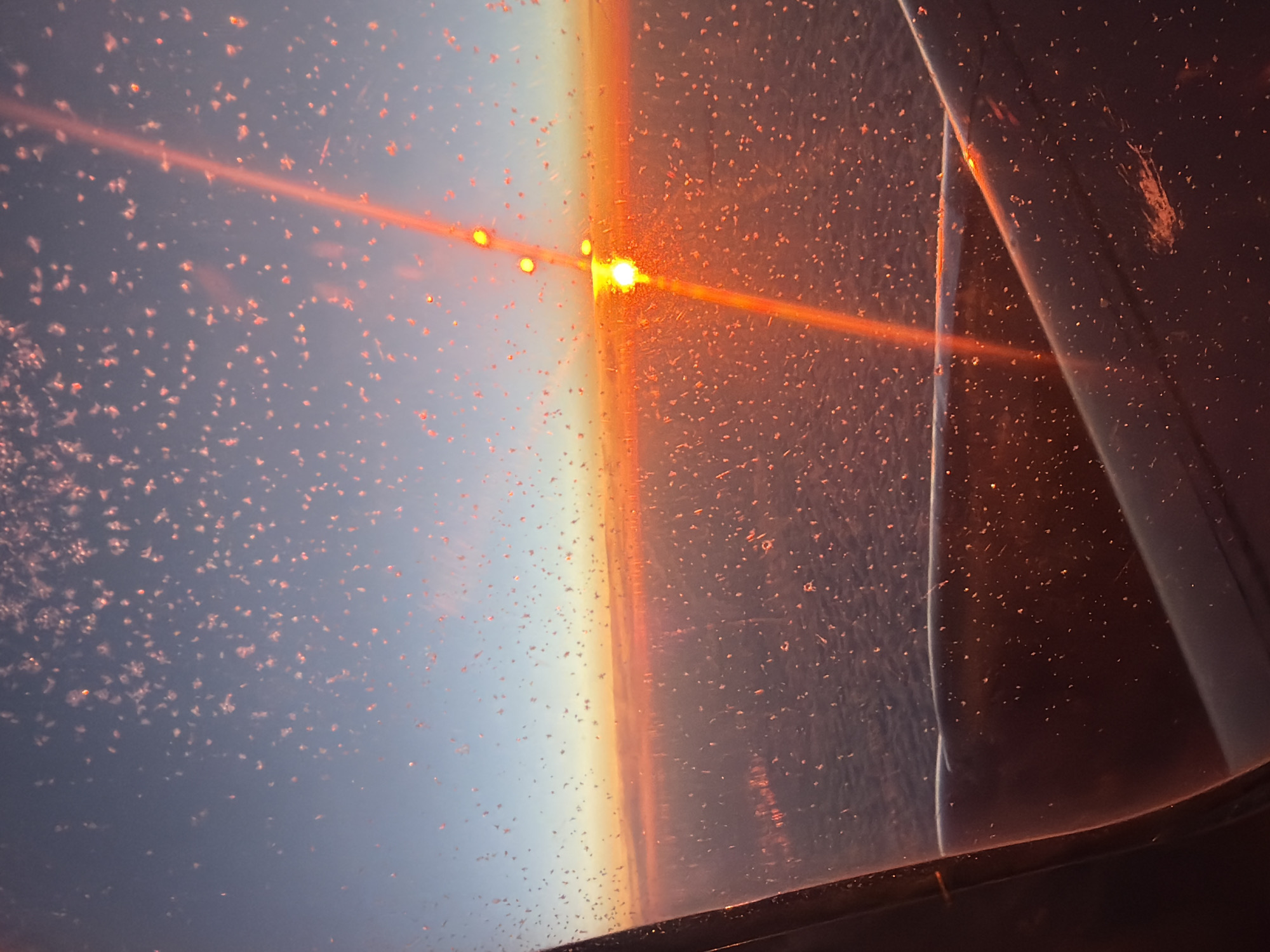The image captures a mesmerizing view from what appears to be an aircraft or spacecraft window, looking towards a brilliant sun at either sunrise or sunset. The dominant feature is a powerful, orange-yellow light source positioned towards the upper middle of the frame, emitting a strong horizontal light streak that extends across the image. This light creates a stark horizon line, transitioning from a warm, fiery orange near the light source to a soft, pastel blue and purple sky on the left.
The window surface itself is covered with countless small, sparkling particles, likely ice crystals or frost, which catch and scatter the intense sunlight, appearing as bright, illuminated specks across the entire view. These particles contribute to the ethereal quality of the scene. On the right side of the image, parts of the window frame or interior structure are visible as dark, curved elements, reflecting some of the ambient light and adding depth to the foreground. The distinct curvature of the horizon suggests a high-altitude perspective. The overall impression is one of immense scale and natural beauty, highlighting the atmospheric conditions and the transition of light at the edge of space or Earth's upper atmosphere. There is no visible text in the image.