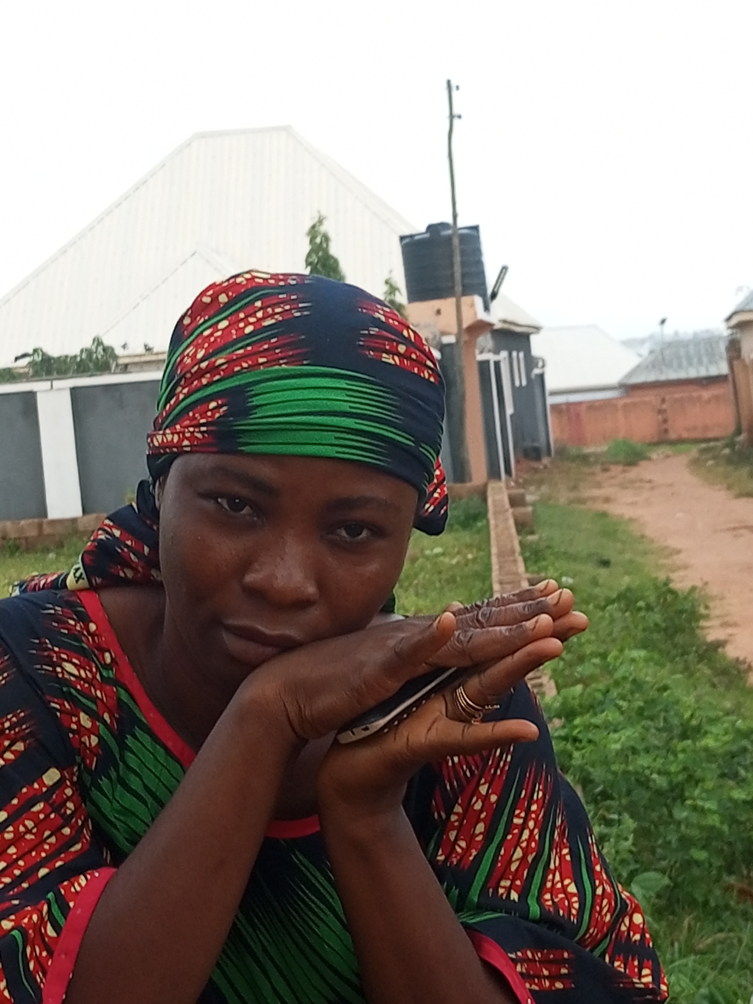 A medium shot photograph captures a woman in the foreground, positioned slightly off-center to the left. She is wearing a dark blue, red, and green patterned headscarf and a matching patterned dress. Her gaze is directed towards the camera, with her head resting on her hands, which are positioned in front of her face. One hand is partially visible, displaying two gold rings on a finger. A smartphone is held between her hands, with its screen facing away from the viewer. The background depicts a somewhat rural or suburban environment, characterized by a dirt path leading into the distance, flanked by green vegetation and a few buildings. A white, sloped roof structure is visible behind the woman, and a black water tank sits atop a building to the right. The sky above is overcast, suggesting a cloudy day. The scene appears to be set in Tempure, Nigeria.
