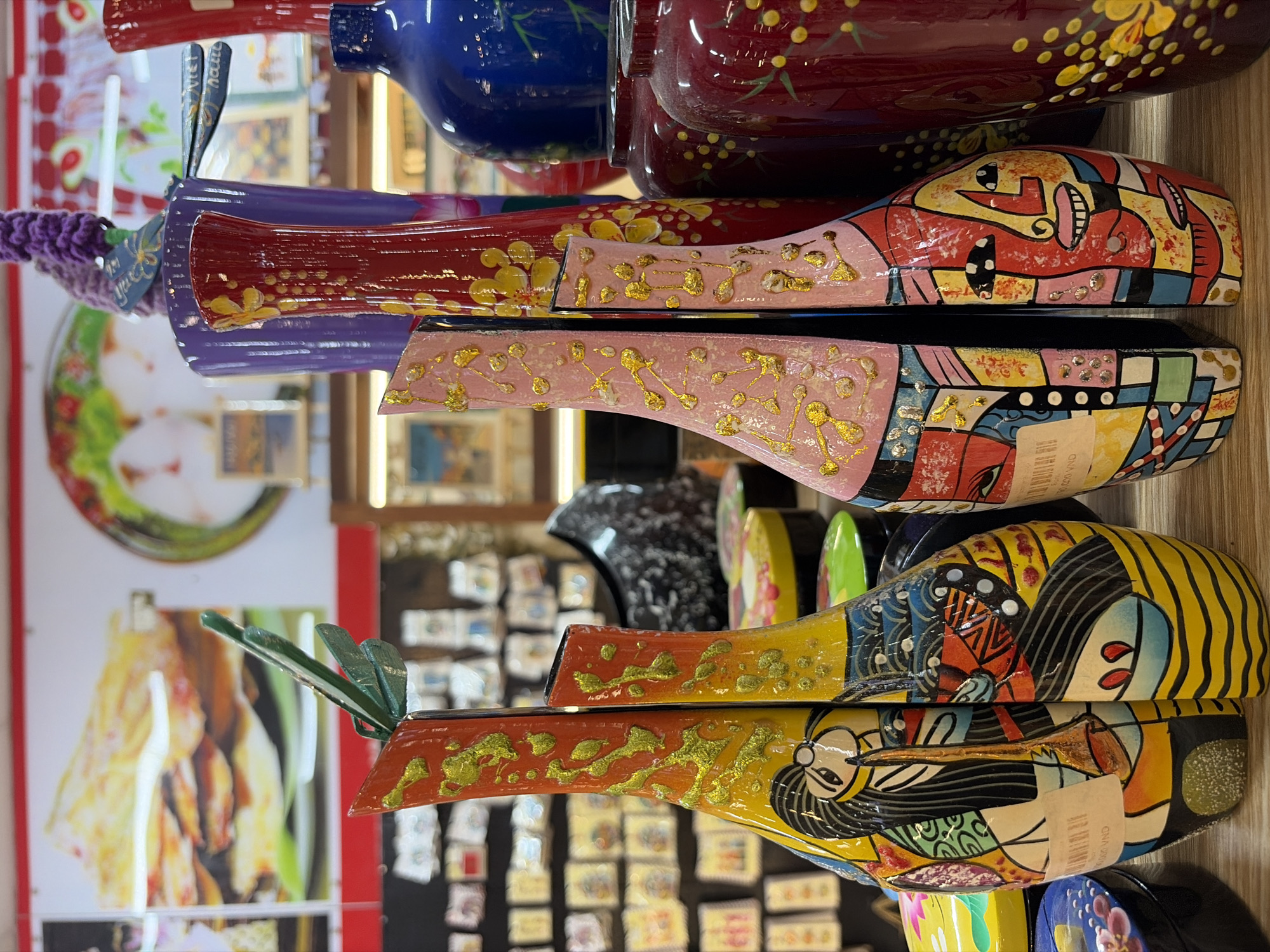 The image displays a close-up view of several colorful, decorative vases and other ceramic items arranged on a wooden surface, likely in a shop or market stall. The vases are intricately painted with vibrant abstract designs and figurative elements, including faces and what appear to be musical instruments. Some of the vases have a textured, raised gold embellishment. In the background, more ceramic items and posters featuring food items are visible, suggesting a retail environment. The overall impression is one of a lively and artistic display. It is not possible to determine the specific location in Da Nang, Vietnam from the image alone.