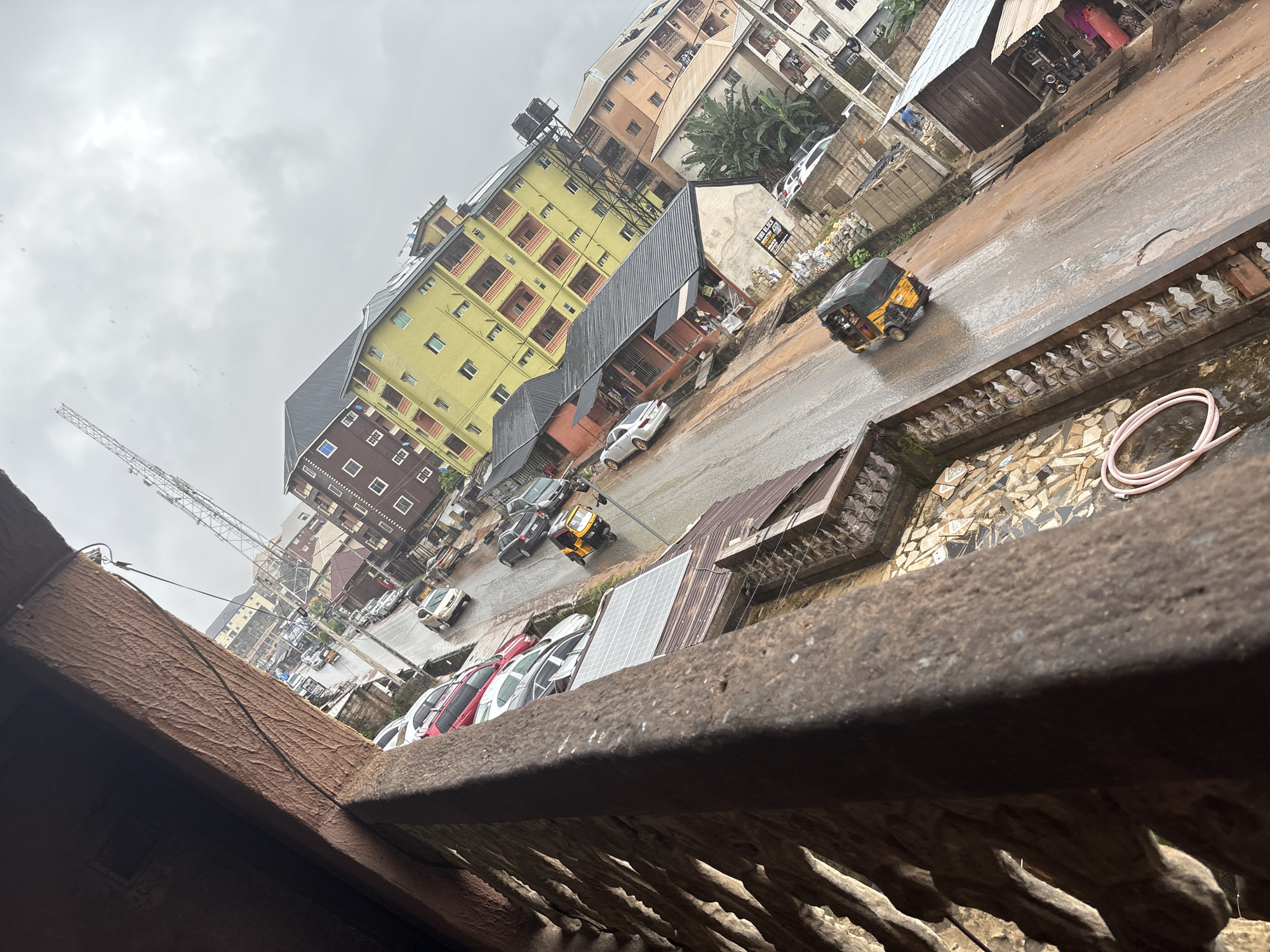 The image depicts a street scene in Nkpor, Nigeria, with a strong tilt to the right. In the foreground, a textured, dark brown railing or balcony structure occupies the left side of the frame, casting a shadow. Beyond the railing, a wet, reddish-brown paved street runs diagonally through the image. Several vehicles are visible on the street, including a yellow and black auto-rickshaw in motion, several cars parked along the side, and other vehicles further down the road.
The buildings lining the street are multi-story structures with a mix of architectural styles. On the right, a prominent yellow building with red balconies stands out, while adjacent to it are buildings of varying heights and colors, some appearing to be residential or commercial. The sky is overcast and grey, suggesting it may have rained recently or is about to. Scattered on the roofs of some buildings are solar panels. In the distance, a tall lattice tower, possibly for telecommunications, is visible against the cloudy sky. The overall impression is of a busy urban environment, likely a market area or a residential and commercial district in Nkpor.