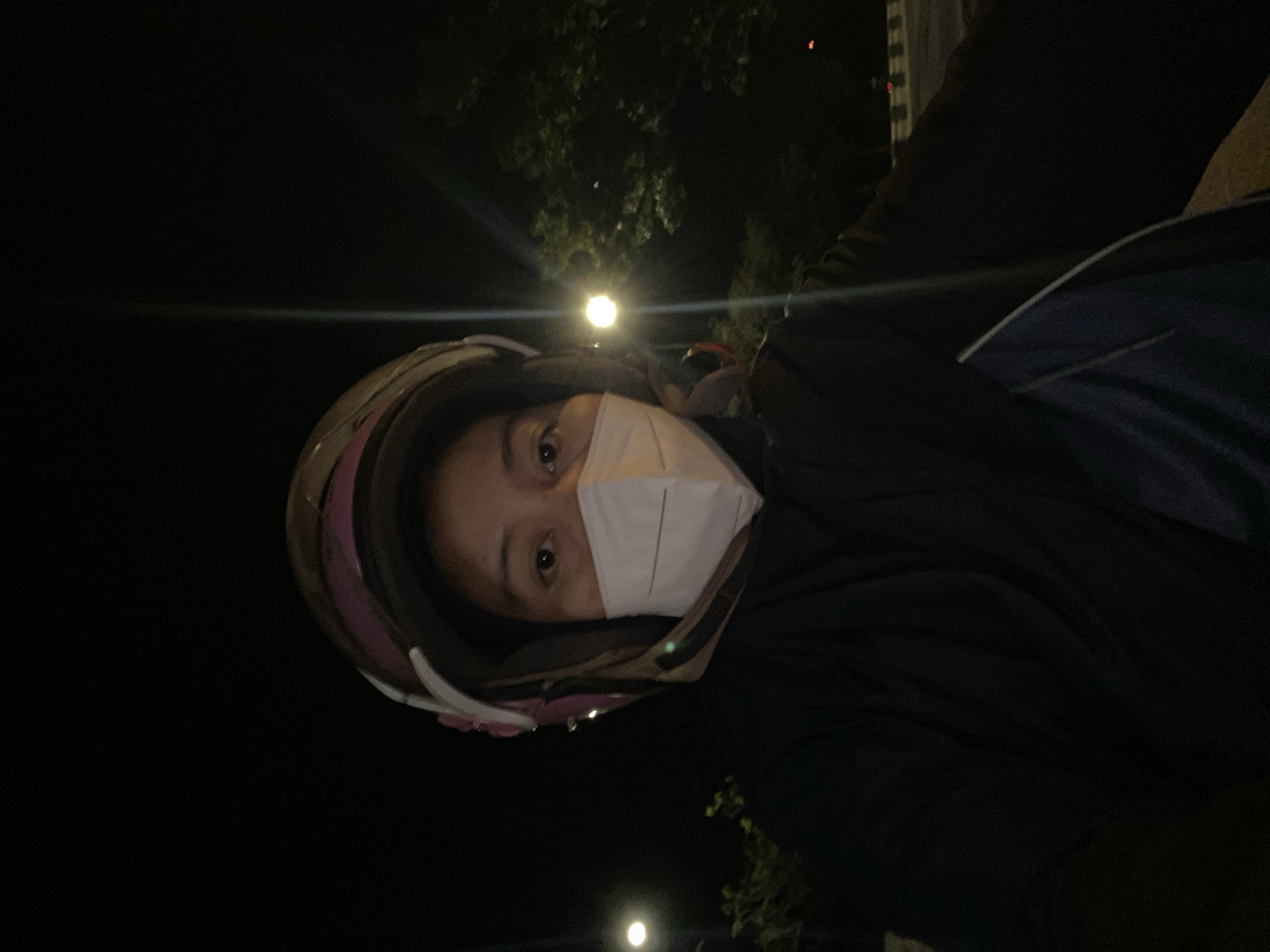 A selfie taken at night shows a person wearing a white mask and a dark helmet with pink and white accents. The person is looking directly at the camera. They are wearing a dark jacket with a blue accent on the sleeve. The background is dark with some trees and lights visible, suggesting an outdoor, possibly urban or suburban, environment. The image is grainy due to low light conditions. The location is specified as Gambut Barat, Indonesia.