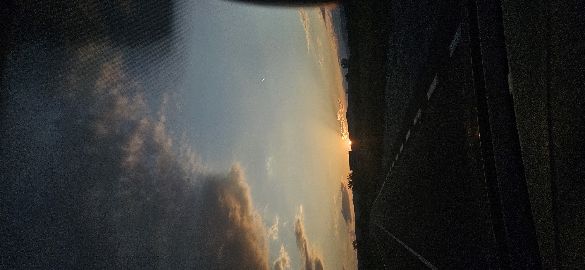This image depicts a sunset over a rural or semi-rural landscape in the vicinity of Dolna Banya, Bulgaria, viewed from the interior of a moving vehicle. The scene is bisected horizontally by the horizon line, with the sky occupying the upper two-thirds and the road and vehicle interior the lower third.
The sky exhibits a dramatic transition from dark, heavy cumulonimbus clouds in the upper left to a clear, pale blue expanse in the upper right. Scattered across the middle of the sky are altocumulus clouds, illuminated by the setting sun with hues of orange, pink, and gold. A distinct, thin white contrail from an aircraft is visible in the clear blue portion of the sky in the upper right. The sun is prominently featured near the horizon in the mid-left, partially obscured by distant landforms, emitting bright orange-yellow light and distinct crepuscular rays that fan upwards.
The horizon consists of a continuous line of silhouetted mountains or hills, appearing dark against the bright sky. Indistinguishable structures or dense vegetation are silhouetted directly beneath the sun.
The foreground is dominated by a dark asphalt road extending from the bottom center into the distance. It features a solid white line on its left edge and broken white dashed lines marking the right lane. The immediate right side of the road is dark and indistinct, suggesting undeveloped land or sparse vegetation. The bottom section of the image shows the dark, underexposed interior of a vehicle, likely the dashboard and a portion of the windshield frame. A visible grid pattern, characteristic of a heated windshield or defroster lines, is present in the upper right quadrant, indicating the photo was taken through glass. No people are visible in the image.
