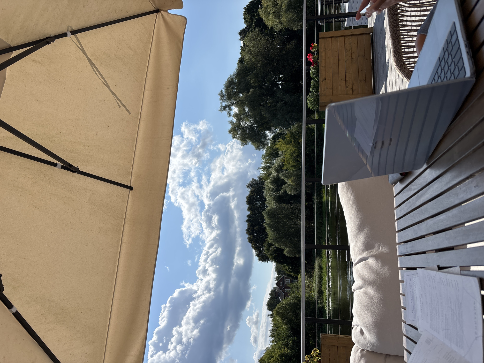 In Zvenigorod, Russia, an outdoor seating area, likely a cafe or restaurant terrace, is depicted under a large, light-colored umbrella. A laptop is open on a wooden slatted table, with some papers in front of it. To the left, a white cushioned sofa is visible. Beyond the terrace railing, a tranquil body of water is seen, reflecting the blue sky and scattered white clouds. The opposite bank is densely populated with lush green trees, and a house with a red roof is partially visible amidst the foliage. Wooden planters with red flowers add a touch of color to the terrace. The overall scene suggests a serene outdoor workspace or relaxation spot with a view of nature.