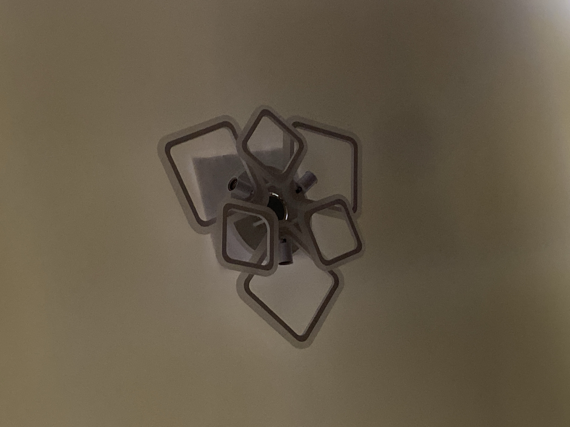 The image displays a ceiling light fixture with a modern, abstract design. The fixture consists of several square-shaped frames arranged in a cluster, emanating from a central point on the ceiling. The frames are outlined in a darker color, possibly brown or black, against a lighter interior. The lighting in the image is dim, casting subtle shadows and giving the fixture a slightly hazy appearance. The ceiling itself is a uniform, pale color, likely white or cream. The perspective is from below, looking directly up at the fixture. There are no people or any other objects visible in the frame. The location is identified as Badalbay, Uzbekistan.