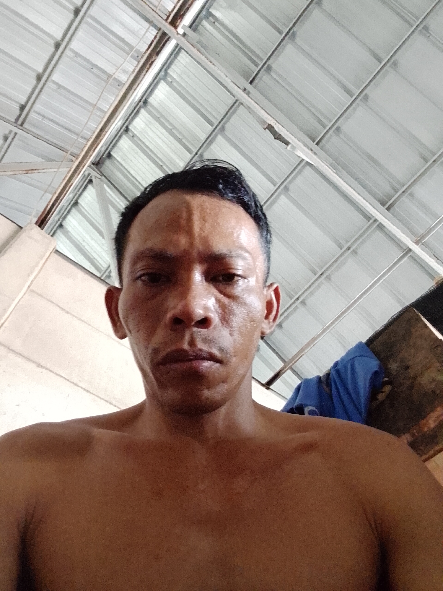 The image shows a shirtless man with short, dark hair, fair skin, and visible sweat on his face and upper body. He is looking directly at the camera with a neutral expression. The background suggests an indoor space, possibly a warehouse or covered outdoor area, with a corrugated metal roof and support beams. A blue cloth is partially visible on the right side. The location is Bukit Ketok, Indonesia.