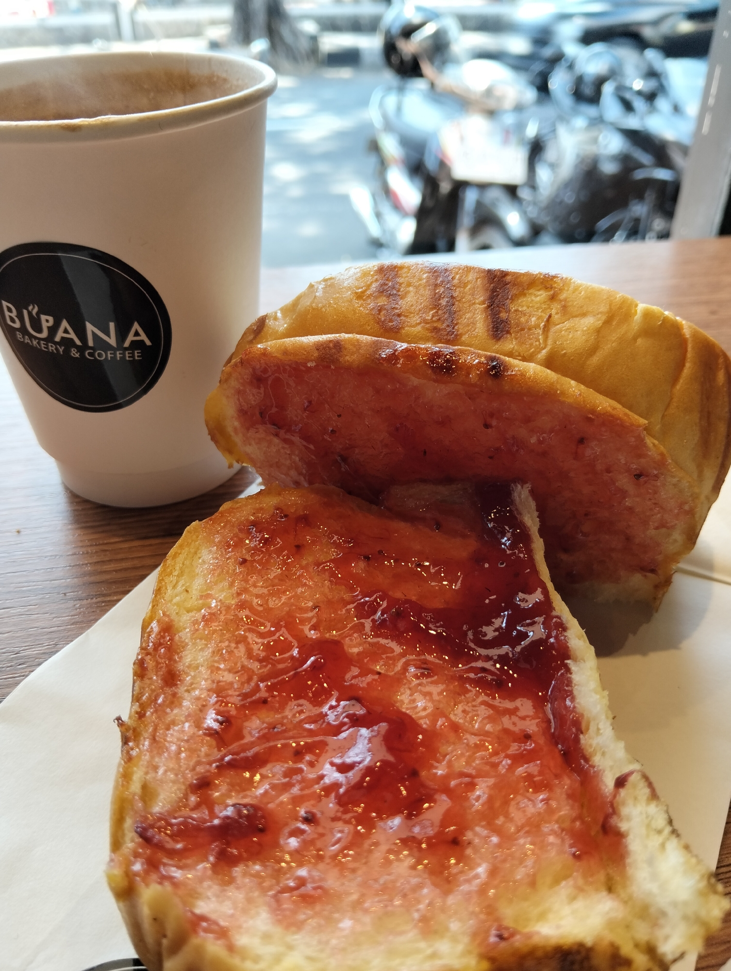 The image depicts a close-up of a white paper coffee cup with the "BUANA BAKERY & COFFEE" logo and two pieces of toast with jam on a wooden table, set against a blurry outdoor background of a street with parked motorcycles and buildings. The toast appears to be toasted with grill marks, and the jam is a vibrant red. The coffee cup is partially filled with a light brown beverage, and steam is visible rising from it. The scene suggests a casual breakfast or snack at a cafe. The location is identified as Magelang, Indonesia.