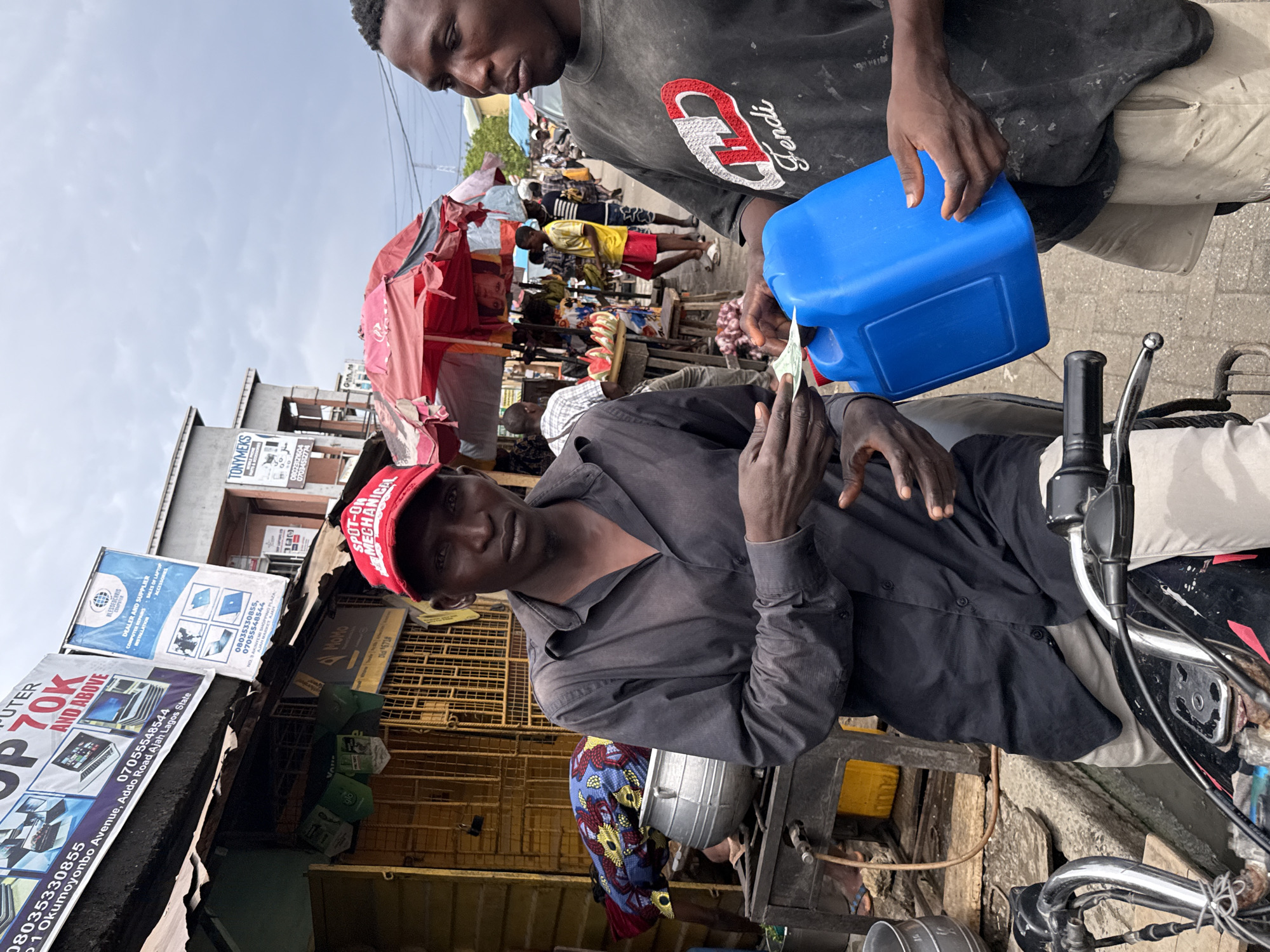 The scene is set in a busy marketplace in Lagos, Nigeria. A man wearing a red cap and a dark gray shirt is seated on a motorcycle, facing the camera. He is holding a wad of Nigerian naira. Another man, wearing a gray shirt with a "Fendi" logo, is pouring fuel from a blue plastic container into the motorcycle's tank. The background reveals a crowded marketplace with stalls shaded by colorful umbrellas. There are commercial buildings with signs advertising computer services and other businesses, including "BLESSEDCHRIS COMPUTER" and "TONYMEKS," with phone numbers and the address "21 Okumoyonbo Avenue, Addo Road Ajah Lagos State." The sky is overcast.