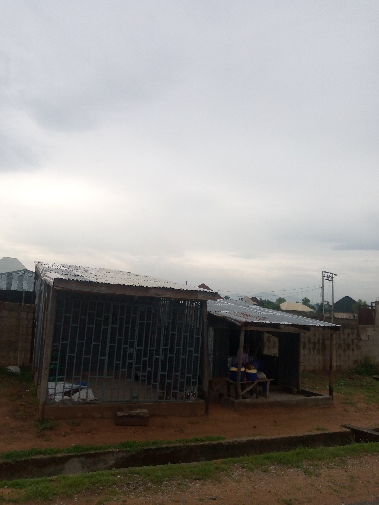 The image shows a street scene in Jalingo, Nigeria, under an overcast sky. In the foreground, there are two small structures with corrugated metal roofs. The structure on the left is larger and enclosed with metal bars, resembling a cage or a secure enclosure. Inside, there appear to be some white tarpaulins. The structure on the right is open on one side and has a person inside, partially obscured. This structure has a wooden frame and corrugated metal siding. Two blue barrels with yellow tops are visible on a wooden bench inside this open structure. The ground is dirt and there is a concrete drainage ditch in front of the structures. In the background, several buildings with metal roofs are visible, as well as some vegetation and a utility pole with wires. The overall impression is of a modest, possibly commercial or residential area.