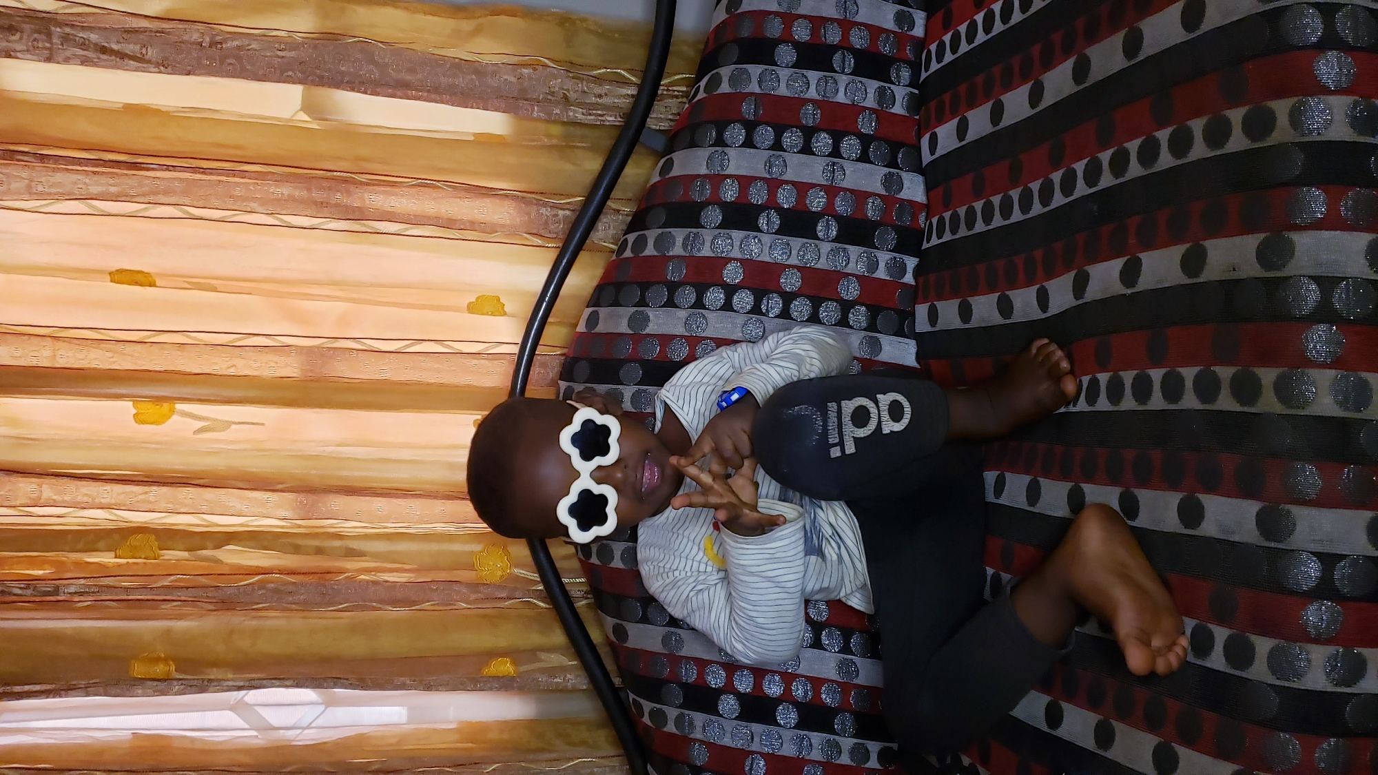 A child wearing flower-shaped sunglasses lies on a striped cushion, with their head near a floral-patterned window covering. The child is also wearing an Adidas garment. The location is Narayi, Nigeria.