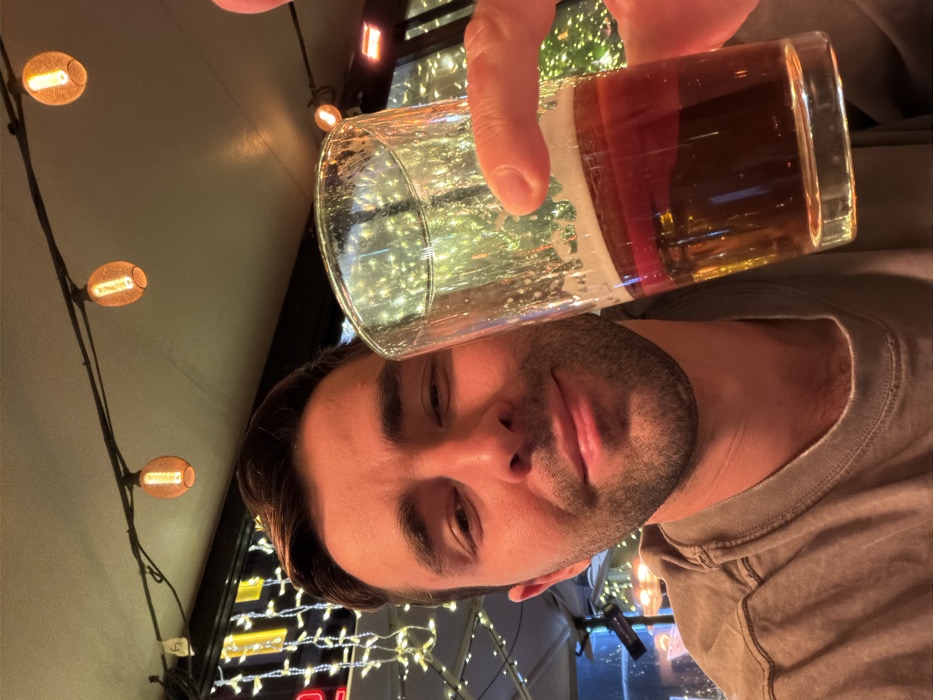 A man holds a pint glass of beer over his face. The glass is mostly clear with some foam and amber liquid. He is wearing a gray t-shirt. A string of lights hangs in the background, along with an illuminated sign. The overall color scheme is warm and slightly muted. The image is rotated to the side.
