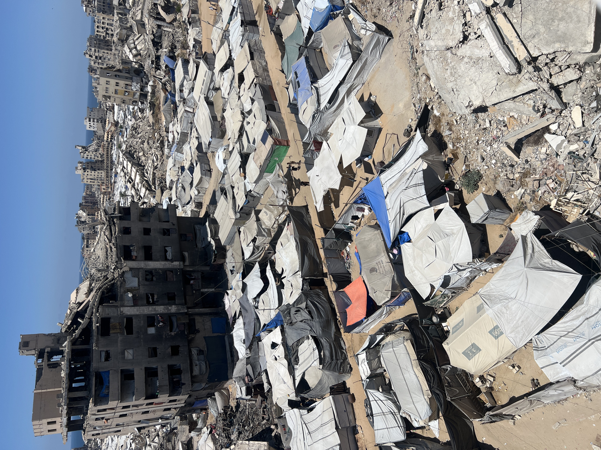 This is an aerial view of a city in Gaza, Palestinian Territories, characterized by widespread destruction and a makeshift settlement of tents. In the foreground, a large, partially destroyed multi-story building is visible, with charred sections and exposed rebar. Surrounding this building and extending into the background is a dense collection of tents, varying in size and color, suggesting a temporary shelter area for displaced individuals. The ground is covered in rubble, debris, and sand. In the mid-ground and background, numerous other damaged buildings are scattered across the urban landscape, with some appearing severely collapsed. The sea is visible in the distance, under a clear blue sky. There are a few people walking among the tents.