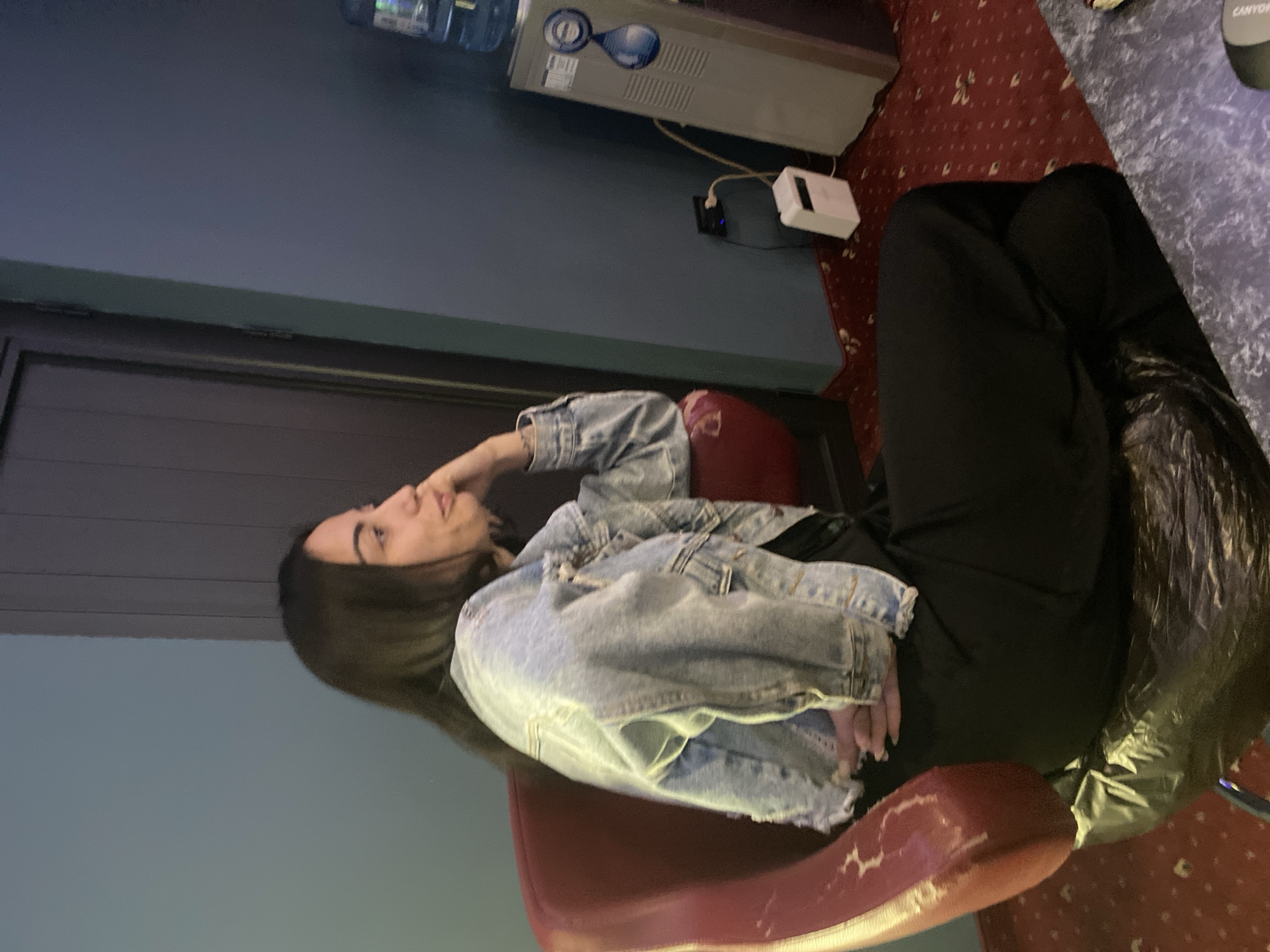 A young woman with long, dark hair is sitting in a red armchair, looking to her right. She is wearing a distressed denim jacket over a black outfit. The armchair is situated in a room with blue walls, a dark wooden door, and a patterned red carpet. To the right of the woman, a water cooler is visible, with an electrical outlet and a small white device nearby. The overall lighting suggests an indoor setting, possibly a lounge or waiting area, with no specific indicators of the city of Beloslav, Bulgaria present in the image.
