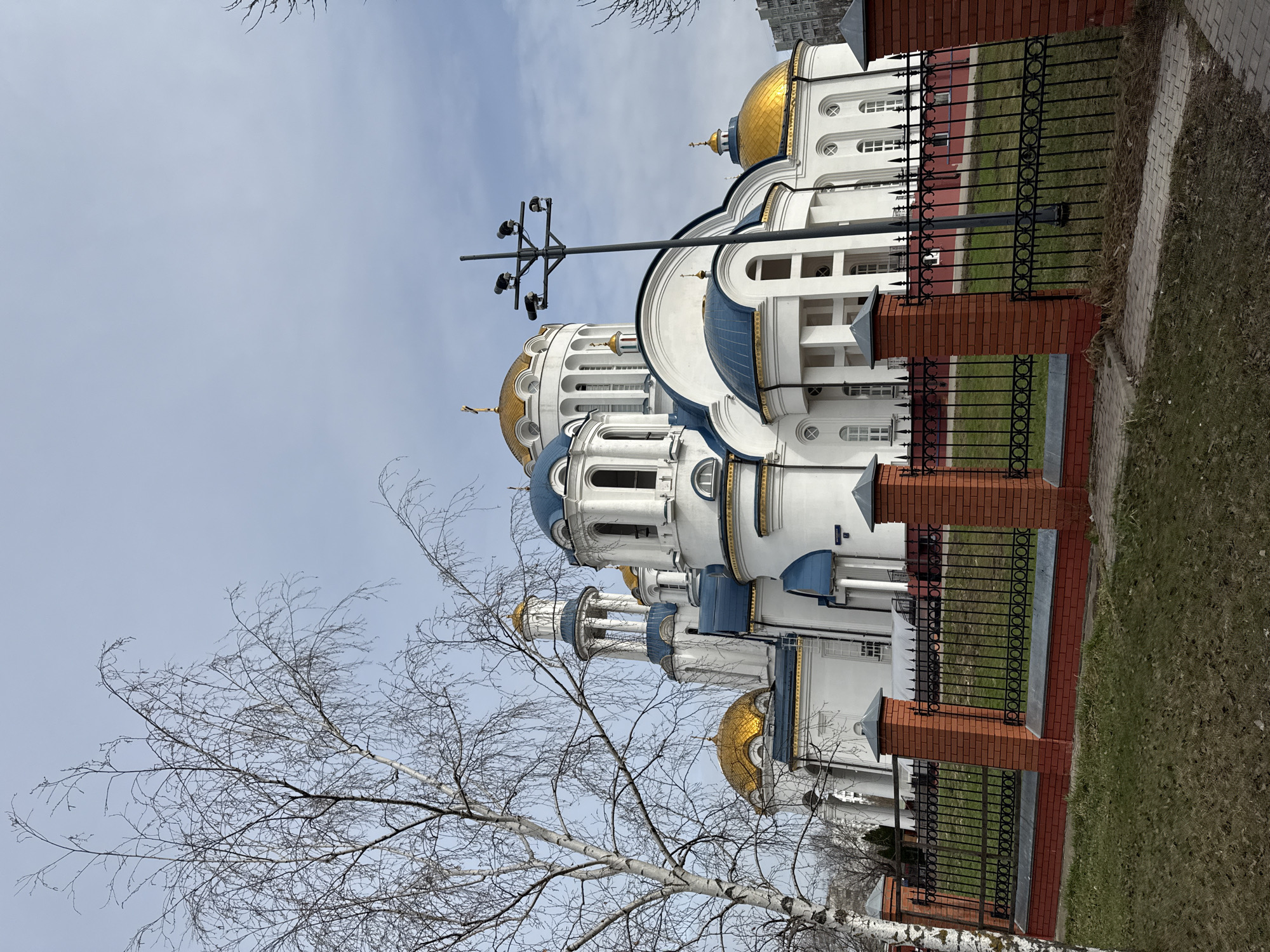 The image captures a white church with blue and gold domes, enclosed by a black metal fence with brick pillars. The church is located in Moscow, Russia. A leafless tree stands in the foreground. The sky is overcast. A light post is visible behind the church.