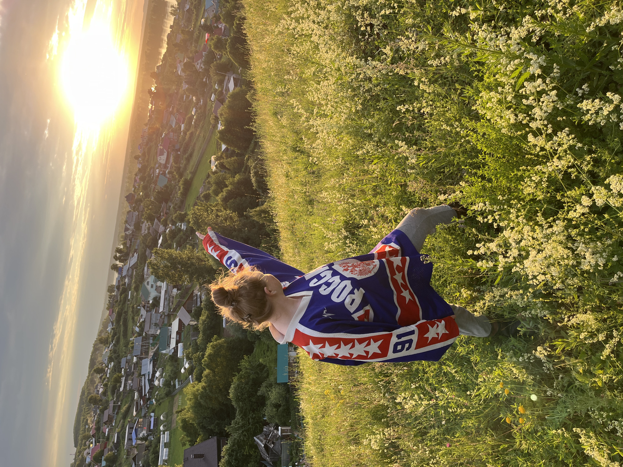 A young person with their hair pulled up in a bun is standing on a grassy hillside overlooking a village at sunset. They are wearing a blue hockey jersey with red and white accents, including stars and the word "РОССИЯ" (Russia) in white lettering. The jersey also has the number "16" on the sleeve. The person is holding up the jersey, which is flowing behind them like a cape. The sun is setting in the background, casting a warm glow over the scene. The village below is composed of small houses with red and gray roofs, surrounded by trees. The hillside is covered in green grass and small white wildflowers. The overall mood is one of freedom and national pride. The time of day is sunset. The location is Olkhovka, Russia.
