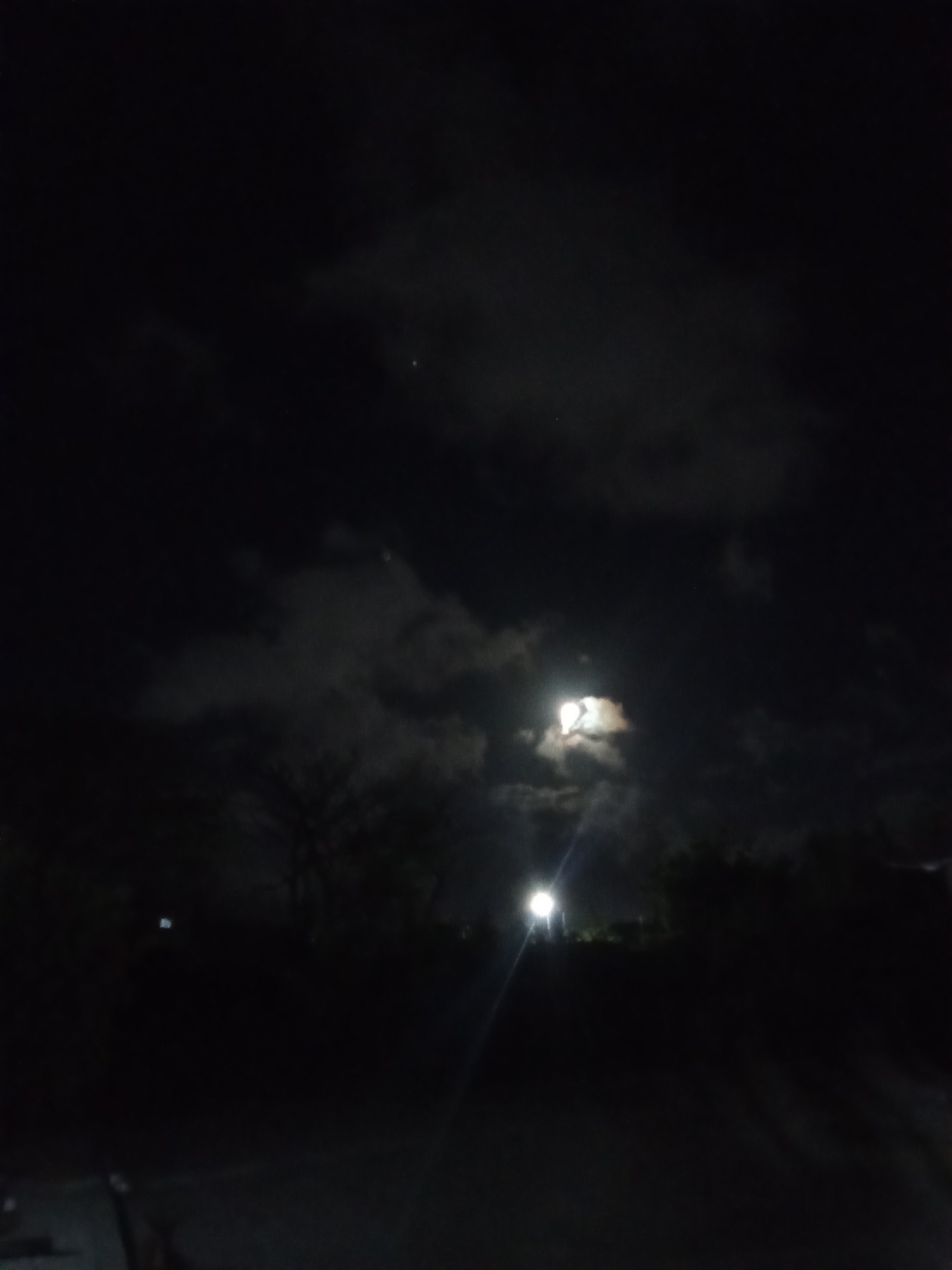 A night scene shows a dark sky with scattered clouds, partially obscuring a bright moon. Below the horizon line, silhouetted trees and indistinct foliage are visible. A light source, possibly a streetlamp, casts a vertical beam upwards, positioned slightly to the right of the moon in the image's composition. The ground is dark and undifferentiated, appearing to be a road or paved area. No people are visible. The location is implied to be Juan Griego, Venezuela.