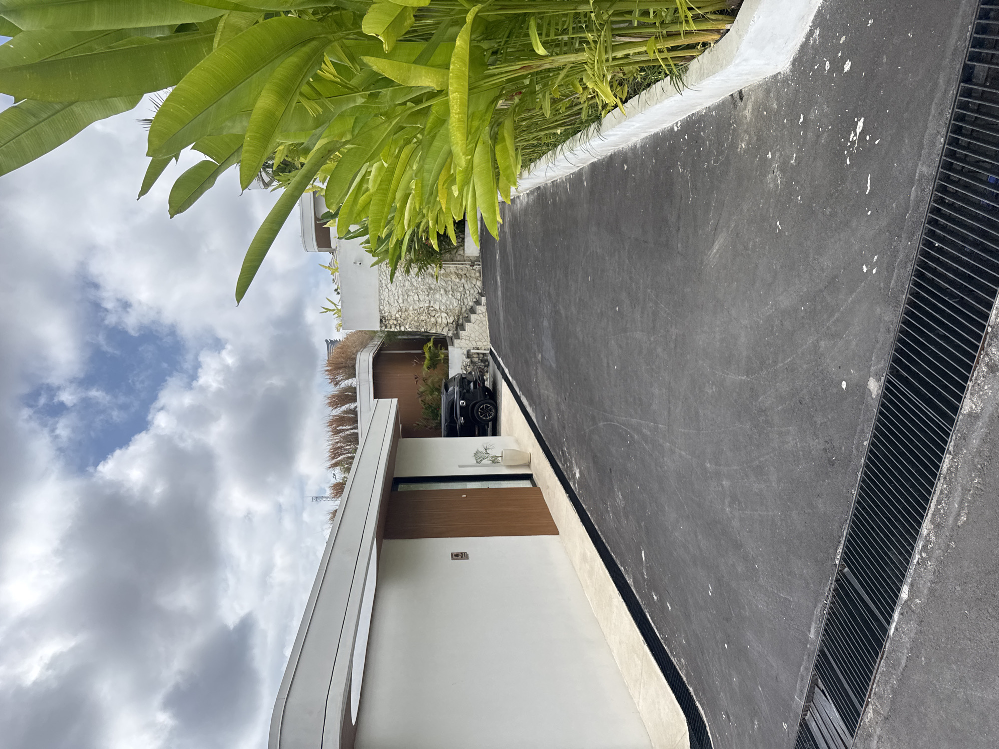 In Kuta Selatan, Indonesia, a paved driveway leads past a modern white building with a wooden door and a parked black SUV. To the right of the driveway, lush green tropical plants with large leaves grow alongside a white concrete planter box. The sky above is partly cloudy with a mix of white and gray clouds.