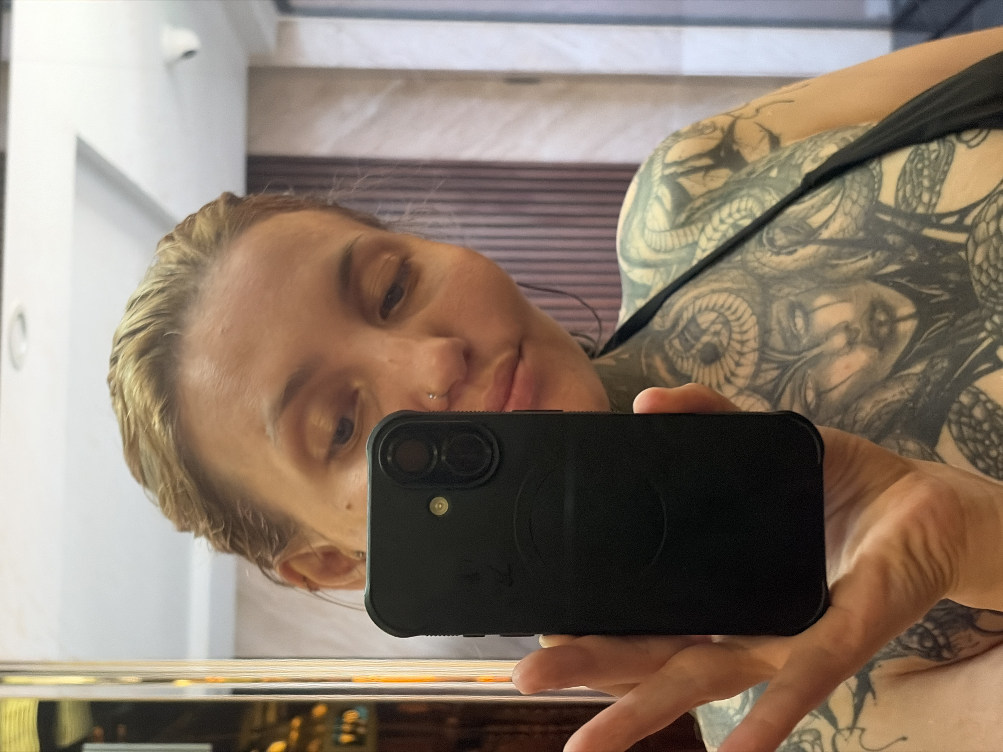 The image is a mirror selfie, presented with a 90-degree clockwise rotation, depicting a person's upper body and face. The subject is a woman with blonde or light brown hair that appears damp or styled back. She has a small nose stud piercing on her left nostril and looks towards the smartphone with a neutral to slightly contemplative expression.
Her shoulders, chest, and upper arm are extensively covered with intricate tattoos. These designs include detailed representations of snakes, scales, and what appear to be faces or demonic figures with horns. She is wearing a dark-colored strap or top, possibly part of a swimsuit or tank top.
She holds a black smartphone with two visible camera lenses and a flash, using it to capture her reflection. The background visible in the mirror behind her features white walls and horizontal wooden paneling, suggesting an indoor setting, potentially a bathroom or hotel room. No text is visible in the image. The location context for the photo is Đà Nẵng, Vietnam.