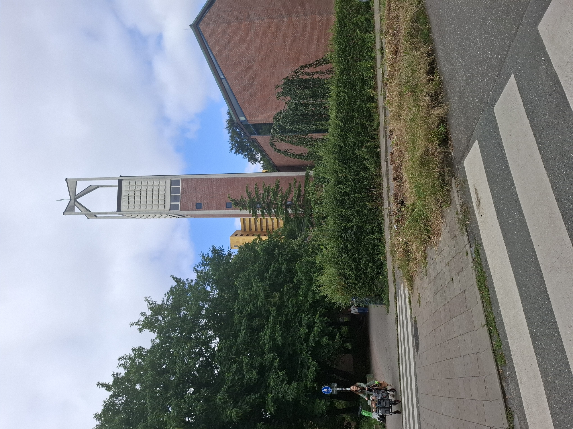 The image shows a view of the St. Bonifatius Church in Hamburg, Germany. In the foreground, a pedestrian crossing is visible on an asphalt road. To the left of the crossing, a woman is pushing a stroller with two children in it along a paved walkway. She is wearing a green jacket and beige pants. A blue sign with a white bicycle symbol is mounted on a pole behind her. Adjacent to the walkway is a dense green hedge.
In the middle ground, the St. Bonifatius Church building is seen. It is constructed of red brick and features a tall, modern bell tower with a distinctive angular structure at its peak. The church has a sloping roof. A weeping tree with branches hanging down is situated in front of the building. Behind the church, a tall yellow building is partially visible.
The background consists of a cloudy sky with some patches of blue. The overall environment appears to be an urban setting with a mix of residential and religious structures, surrounded by greenery.