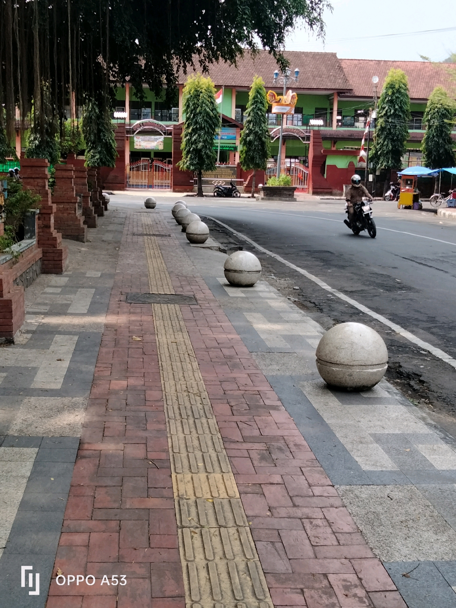 The image shows a street scene in Trenggalek, Indonesia, with a sidewalk in the foreground and a building in the background. On the sidewalk, there are several concrete spheres placed at intervals. The sidewalk is paved with red bricks and has a textured yellow strip, likely for tactile paving. A person on a motorcycle is riding on the street. In the background, a building with green and red walls and a tiled roof is visible, with trees and a red brick wall in front of it. There are also some market stalls or vendors set up on the side of the road. The overall scene depicts a typical urban street environment in Indonesia.