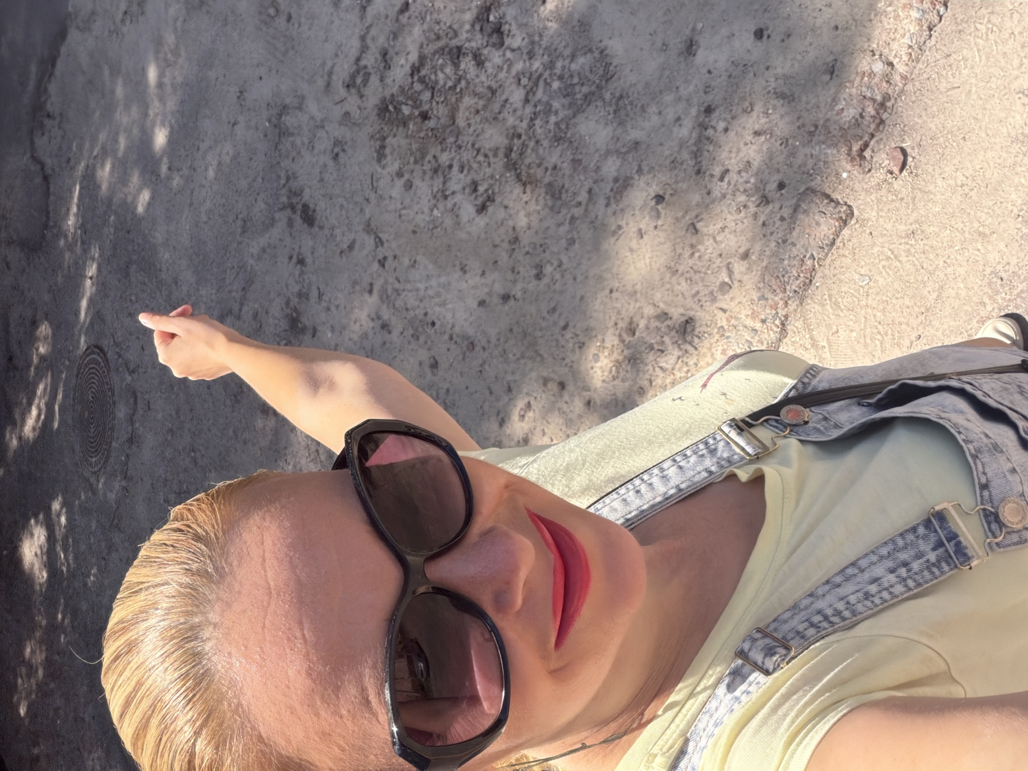 A woman with blonde hair pulled back is taking a selfie outdoors, looking at the camera and smiling. She is wearing large black sunglasses, bright pink lipstick, a pale yellow t-shirt, and light-wash denim overalls. Her right arm is raised, pointing her index finger towards the upper right of the frame. The background consists of a rough, textured ground surface, possibly dirt or gravel, with patches of shadow cast by unseen foliage. A circular metal grate is visible in the upper left corner of the background. The lighting suggests it is a sunny day.