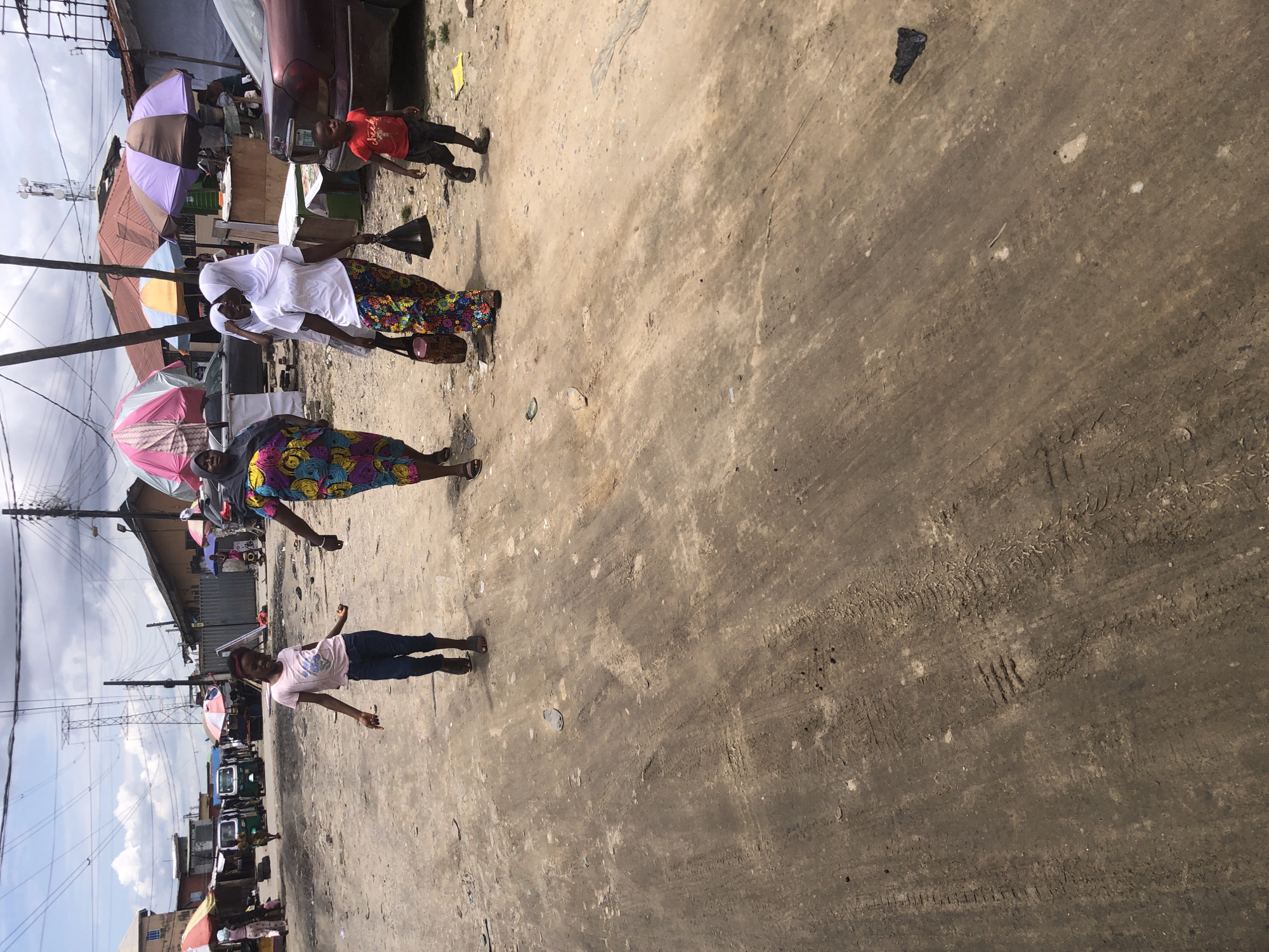 The image depicts a dusty street scene in Ado, Nigeria, likely a market or residential area. The ground is unpaved and shows tire tracks from vehicles. In the foreground, a young girl with dark hair, wearing a pink t-shirt and blue jeans, is walking. To her right, a woman in a colorful dress and headscarf walks, and to her right, another woman in a white headscarf and patterned pants carries a dark bag. A young boy in a red shirt and dark shorts walks ahead of them. In the background, market stalls with umbrellas, buildings, and utility poles are visible, along with a red car and a green and white tricycle-like vehicle. The sky is cloudy.