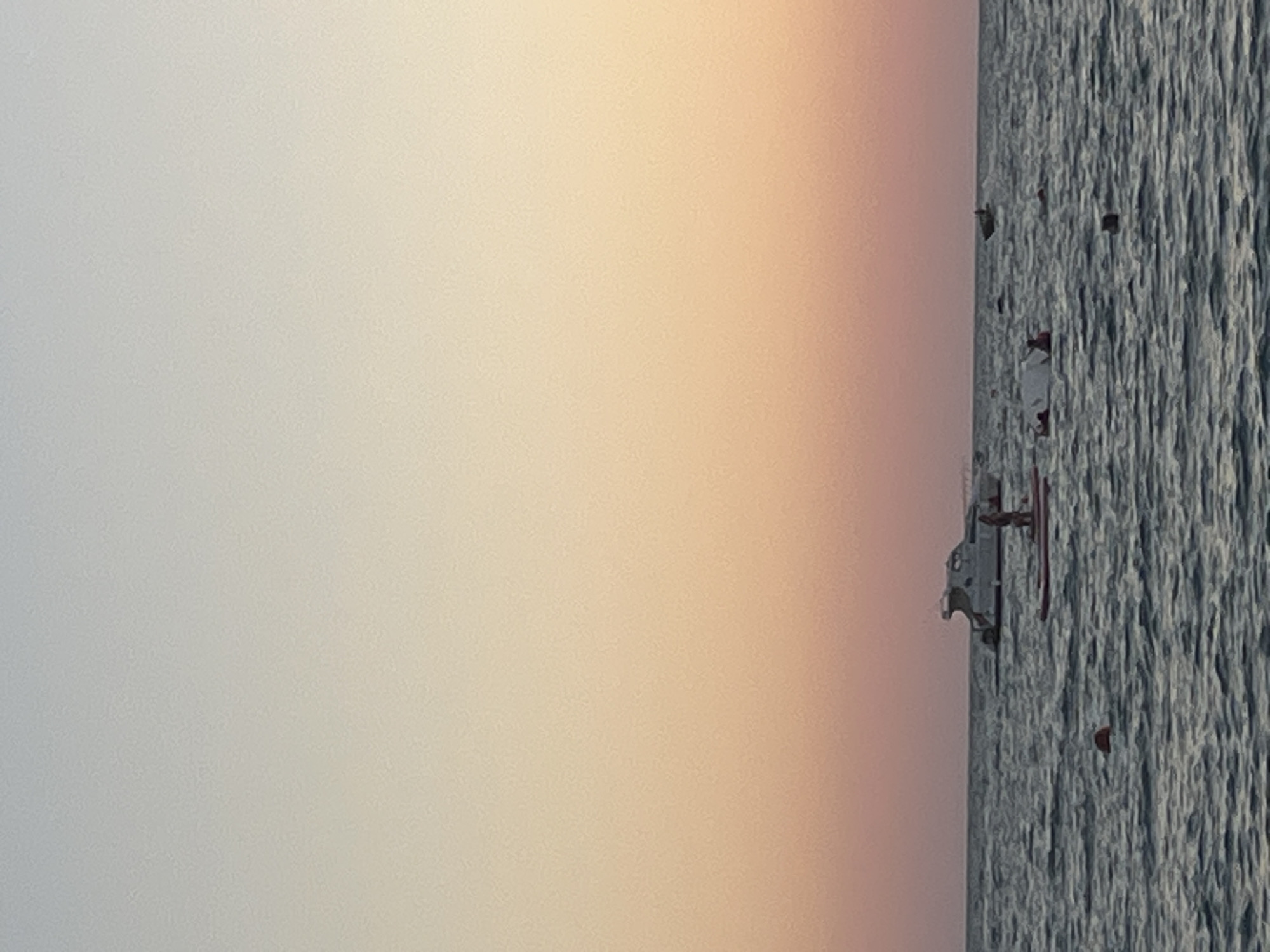 The image depicts a serene seascape at dusk, likely near Sochi, Russia. The sky transitions from a pale blue at the top to a warm gradient of peach and pink towards the horizon, creating a soft, diffused light. The sea is a deep, rippling blue, with gentle waves.
Several objects are scattered across the water. In the mid-ground, a white catamaran-style boat is anchored, its sleek design partially visible. To the right of the boat, a person is standing on a paddleboard. Further out in the distance, a small motorboat is visible. Closer to the foreground, a red buoy floats on the water.
Scattered around the water are several people engaged in recreational activities. Two individuals appear to be on a deflated inflatable structure, while another person is paddling on a red paddleboard. Another person is visible further to the right, appearing to be in the water. The overall impression is one of leisure and a peaceful end to the day. The presence of multiple recreational watercraft and individuals suggests a popular spot for water activities.
