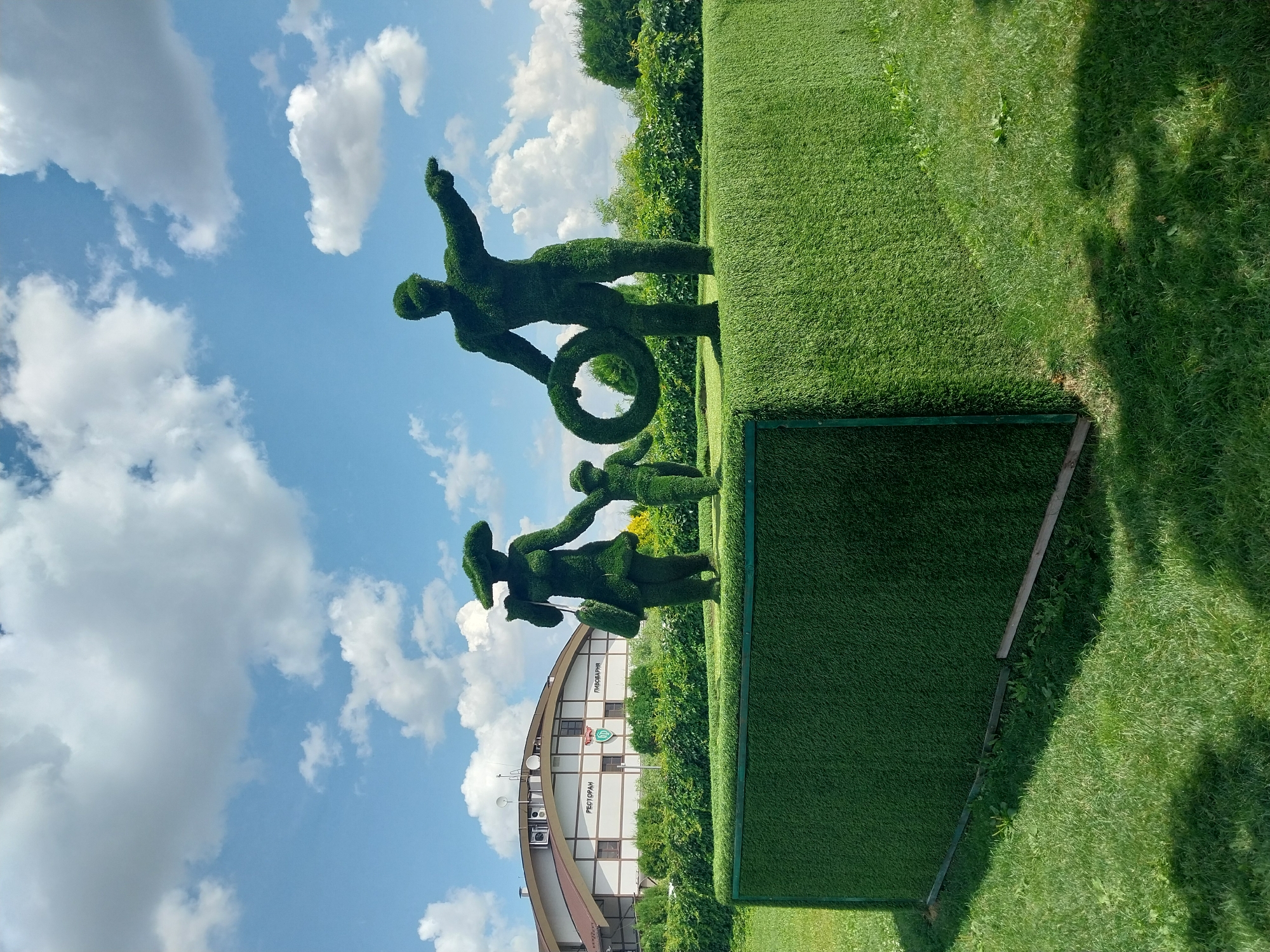 In Dnipro, Ukraine, a topiary sculpture depicts a family of three against a backdrop of a building and a clear blue sky with scattered white clouds. The sculpture is situated on a green, artificial turf platform. The family is composed of a man, a woman, and a child. The man, on the right, is larger than the other two figures and holds a wreath in his left arm, while his right arm is extended, pointing forward. The woman, to the left, is dressed in a hat and carries a purse on her left shoulder. The child stands between the man and woman, holding the woman's hand. Behind the sculptures, a low, green hedge runs horizontally, separating the platform from a building with a brown roof and white and beige siding. Signs on the building read "РЕСТОРАН" (RESTAURANT) and "ПИВОВАРНЯ" (BREWERY). The area in front of the platform is covered in lush green grass, with some shadows cast by an unseen object.