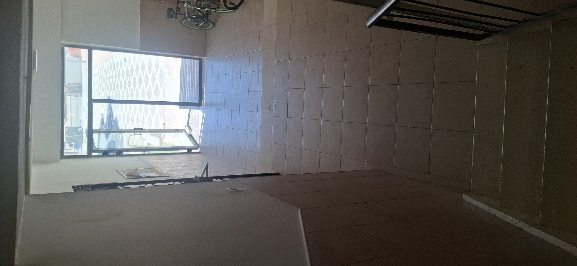 The image depicts the entrance to a building in Yerocham, Israel, likely a residential building or an office space. The perspective is from a staircase looking down towards the entrance doors and the outdoor area beyond.
The interior consists of a tiled floor with large, light-colored square tiles. The walls are paneled with light brown tiles on the lower portion and painted a light color on the upper portion. A metal handrail is visible on the left side of the staircase.
The entrance area features double glass doors, one of which is ajar. Through the doors, a patterned paved outdoor area is visible, with diamond-shaped light and dark gray patterns. A car can be seen parked outside through the glass. On the right side of the entrance, two bicycles are parked. One is green and black, and the other appears to be darker.
The overall impression is a functional, well-lit entrance area, suggesting it is a public or semi-public space. The presence of bicycles indicates that cycling is a common mode of transport in the area. The patterned pavement outside suggests a modern or well-maintained urban environment. The image appears to have been taken during daylight hours, with natural light illuminating the scene.