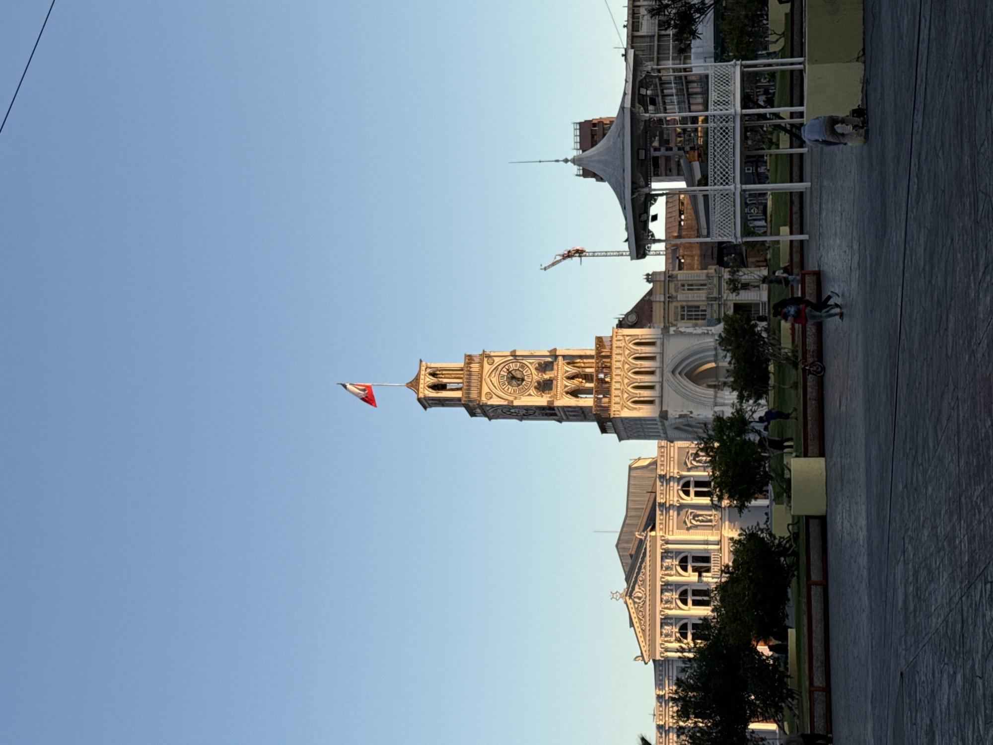 The image depicts the Plaza Añañucas in Iquique, Chile, during a clear, sunny day with a bright blue sky. The plaza features a prominent clock tower with a Chilean flag flying from its peak. To the left of the clock tower is a neoclassical building. In the foreground, a paved plaza area is visible, with several people walking and interacting. A white gazebo with intricate latticework stands to the right of the clock tower. In the background, construction cranes and other buildings are present, indicating ongoing development or urban activity. The overall scene is one of a public space in a city, bathed in the warm light of what appears to be late afternoon or early morning sun.