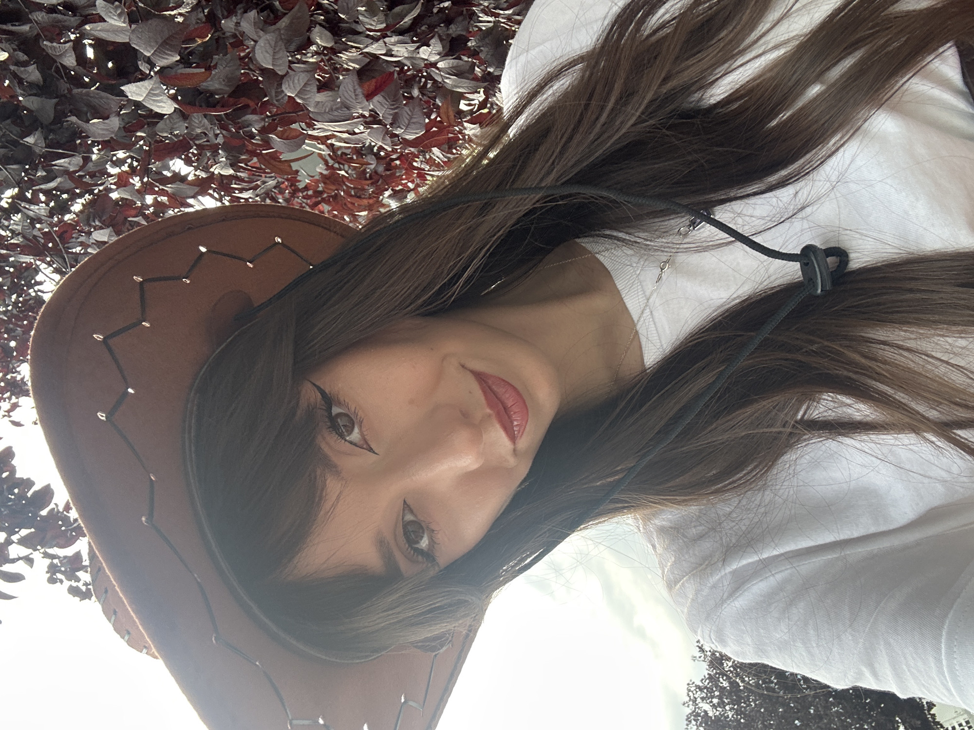 A young woman with long brown hair and bangs, wearing a brown cowboy hat and a white t-shirt, smiles at the camera. She has dark eyeliner and red lipstick. The background consists of dark red leaves on a tree and a bright, overcast sky. The image appears to have been taken outdoors in a garden or park. There is no discernible information in the image to place it in Grocka Urban Municipality, Serbia.