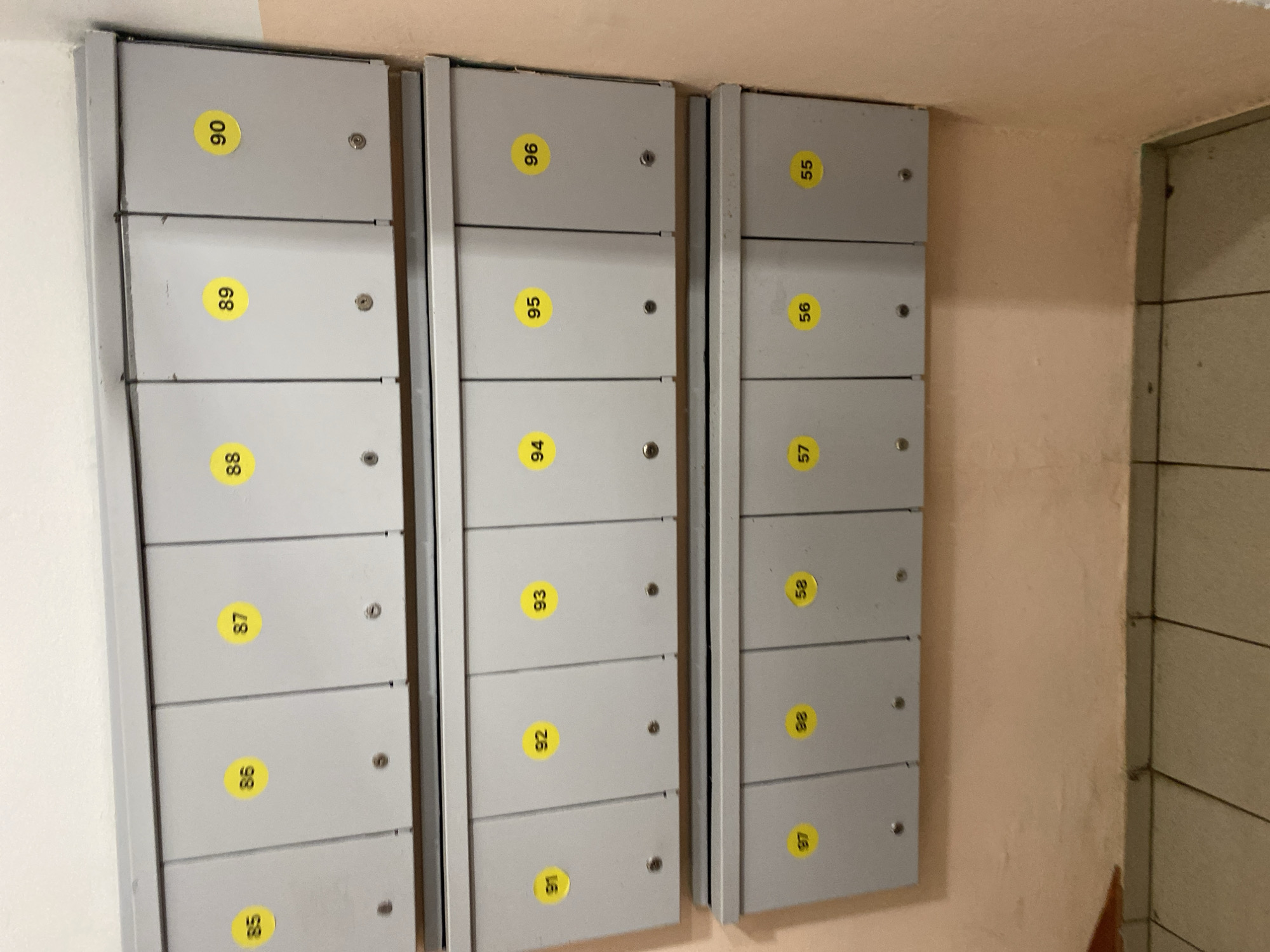 The image shows a wall of metal mailboxes in a building in the city of Khimki, Russia. The mailboxes are arranged in three rows, with each mailbox door marked with a yellow sticker displaying a number. The top row has mailboxes numbered 85, 86, 87, 88, 89, and 90. The middle row has mailboxes numbered 91, 92, 93, 94, 95, and 96. The bottom row has mailboxes numbered 97, 98, 58, 57, 56, and 55. The mailboxes appear to be made of light gray metal and are mounted on a wall painted in a peachy-pink color. The floor in the foreground is tiled with square, light-colored tiles. The lighting suggests an indoor environment, likely a common area within an apartment building or office complex. There are no people or animals visible in the image. The overall scene is one of a functional, but somewhat dated, building interior.