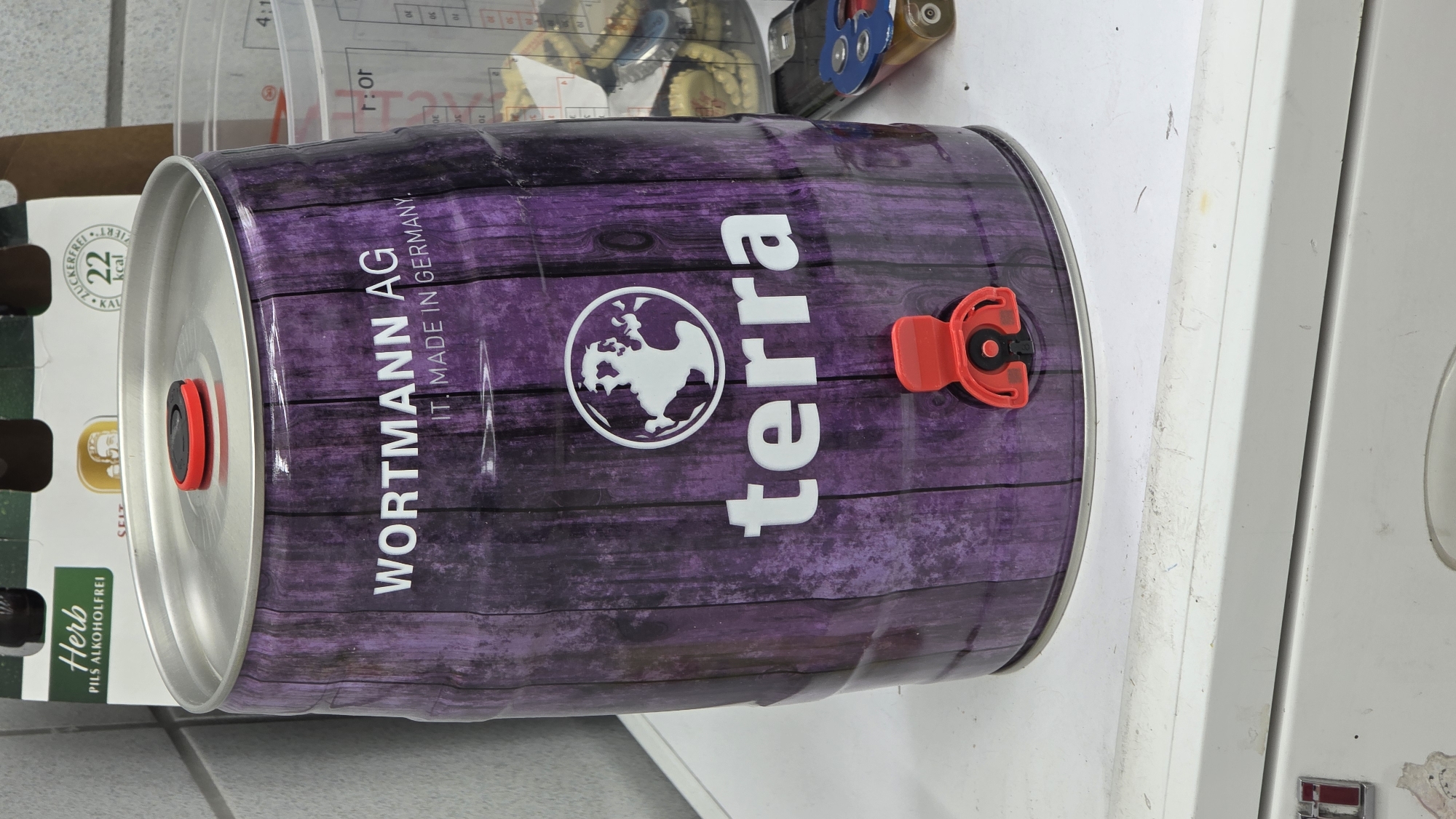 A medium shot shows a purple and black wooden patterned keg with a red tap on a white surface. The keg has white lettering that reads "WORTMANN AG" and "IT. MADE IN GERMANY." Below this, a white logo with a white figure in a circle is visible, followed by the word "terra" in white lettering. The keg has a silver lid with a small red plastic cap. In the background, a clear plastic container filled with bottle caps and a partially visible beer carton are present. To the right of the keg, a black mobile phone and a multi-tool are resting on the white surface. The scene appears to be indoors, possibly in a kitchen or storage area, with a tiled wall visible in the upper left corner.
