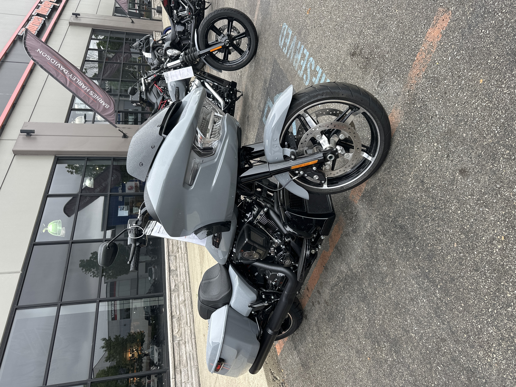 A grey Harley-Davidson motorcycle is parked in front of a building with large windows. The motorcycle is angled towards the viewer, with its front wheel turned slightly to the left. The motorcycle has a large front fairing, saddlebags, and a black exhaust system. In the background, other motorcycles are visible, as well as a banner that reads "BARNES HARLEY-DAVIDSON". The ground is paved with asphalt, and there are orange markings on the ground. The building appears to be a dealership or showroom. The scene is outdoors, and the lighting suggests it is daytime.
