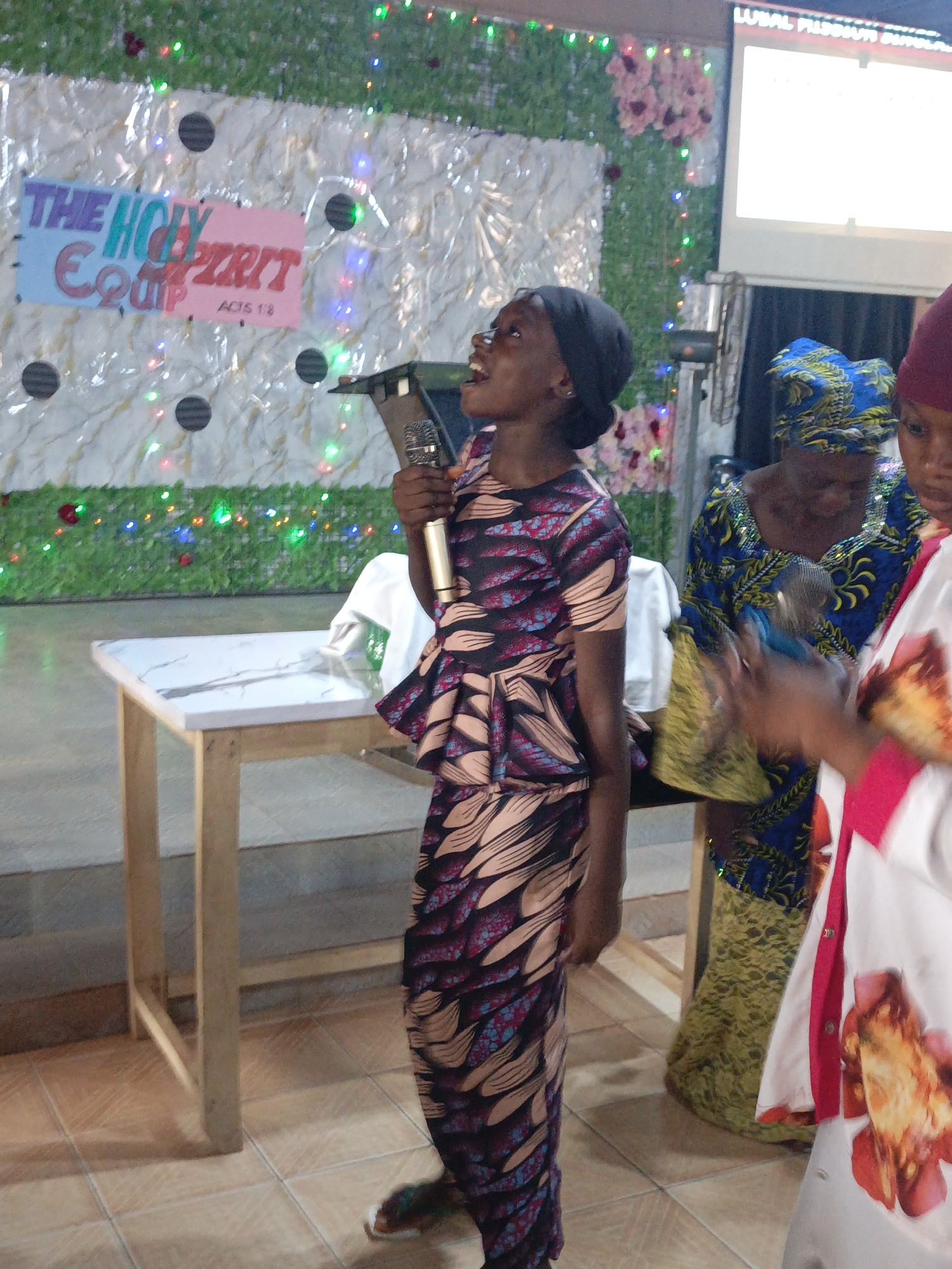 In a brightly lit indoor setting in Akwanga, Nigeria, a young, dark-skinned woman wearing a patterned, peplum-style dress and a black head covering stands at a small table, holding a microphone and singing. Behind her, a backdrop is decorated with green foliage, twinkling lights, and flowers, with a sign reading "THE HOLY SPIRIT EQUIP ACTS 1.8." To her right are two women; one in a blue and yellow patterned dress, holding a microphone, and another partially visible in a white shirt with a red floral pattern. A blank projection screen is visible in the upper right corner. The floor is tiled.