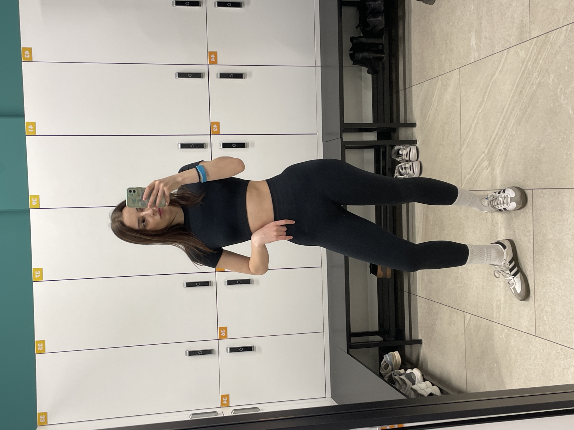A young woman stands in a modern locker room, taking a mirror selfie with her light-colored smartphone. She is dressed in athletic wear, consisting of a black short-sleeved crop top and black form-fitting leggings. A light blue wristband is visible on her left arm. She wears white crew socks and white and black athletic sneakers, which appear to be Adidas Sambas.
The locker room features white, rectangular lockers with dark, minimalist handles or locks. Yellow square labels with black numbers such as "33," "34," "38," "39," "43," "44," and "45" are affixed to the lockers. A dark bench or shelving unit runs along the base of the lockers and extends to the right side of the frame, where several other pairs of sneakers are stored. The floor is covered with large, light-colored tiles that have subtle, wavy patterns. A turquoise-colored wall is visible on the far left, adjacent to the lockers. The woman has long, dark hair and is looking directly into her phone, held up to capture her reflection. The setting, as per context, is in Moscow, Russia.