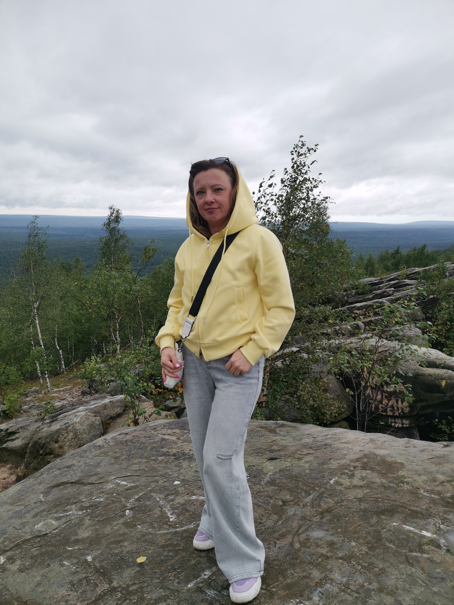 A woman stands on a rocky outcrop overlooking a vast forest under an overcast sky. She is wearing a light yellow hooded jacket, light gray flared jeans, and light purple sneakers. A black strap bag is slung across her body. Her hands are in her pockets. Sunglasses are perched on top of her head. The background shows a panoramic view of a dense forest stretching to the horizon, with several birch trees in the foreground. The rocks in the foreground appear weathered and have some white markings on them. The location is likely the Katchkanar mountain range, known for its unique rock formations and extensive forests, near the city of Gubakha, Russia.