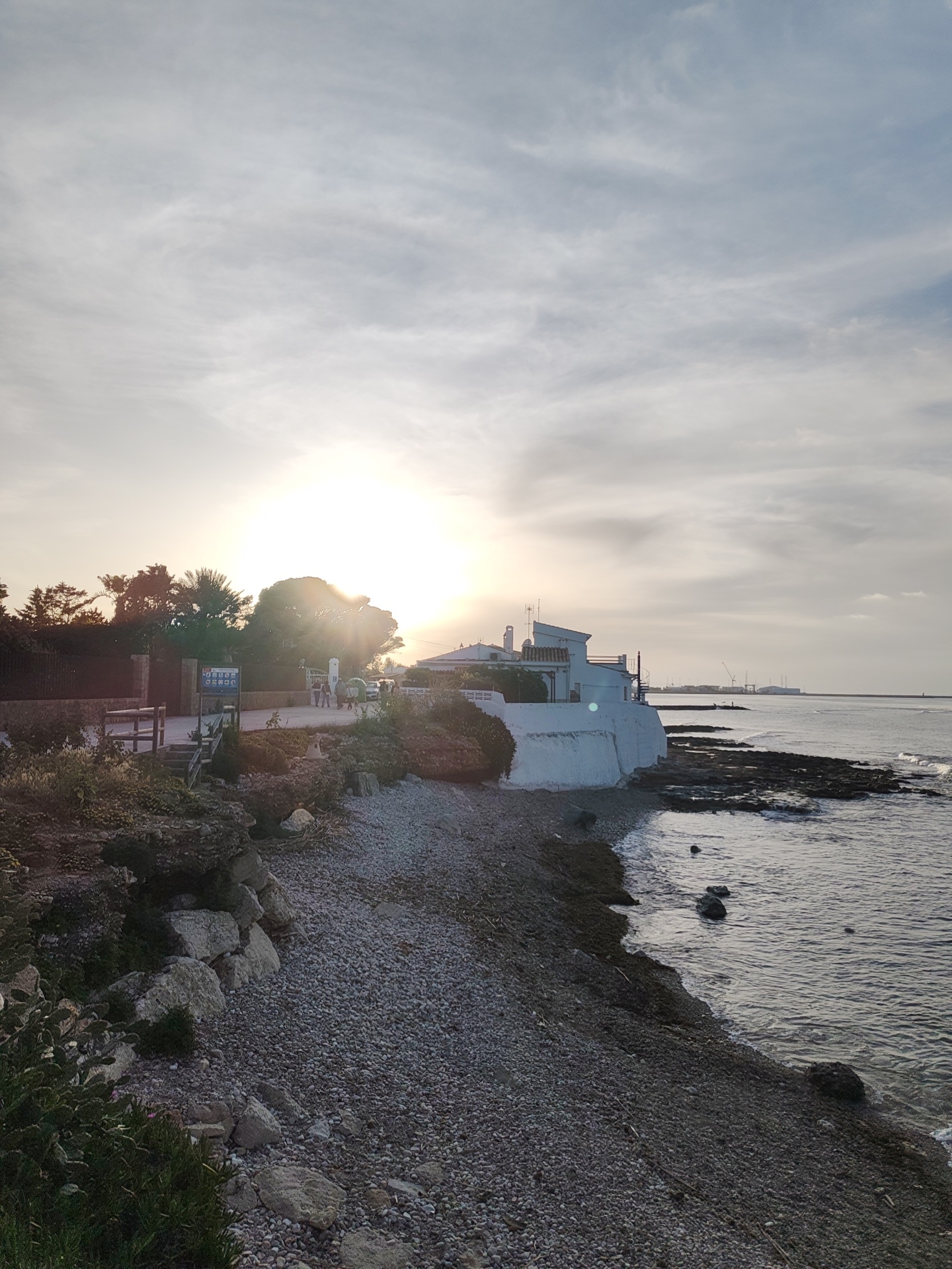 The image depicts a coastline scene in Dénia, Spain, at what appears to be either sunrise or sunset. The sun is positioned behind trees and a building, creating a strong glare. To the right of the frame, a body of water extends towards the horizon, where distant industrial structures are visible. A rocky shoreline is prominent, with small waves lapping at the edge. The shore consists of pebbles. A white building with a visible roof and chimneys stands on the shore. Vegetation is seen on a small cliff and in the foreground, to the left. A sign and wooden steps lead from the vegetation to the pebble beach. People can be seen at the top of the steps.