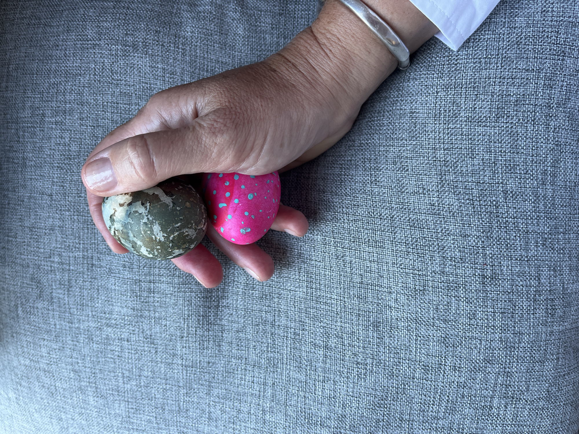 A person's hand, wearing a silver bracelet and a white shirt cuff, holds two decorative eggs against a gray woven fabric background. One egg is pink with blue dots, while the other is mottled with shades of green, gray, and white. No specific location in Denpasar, Indonesia is discernible from the image.