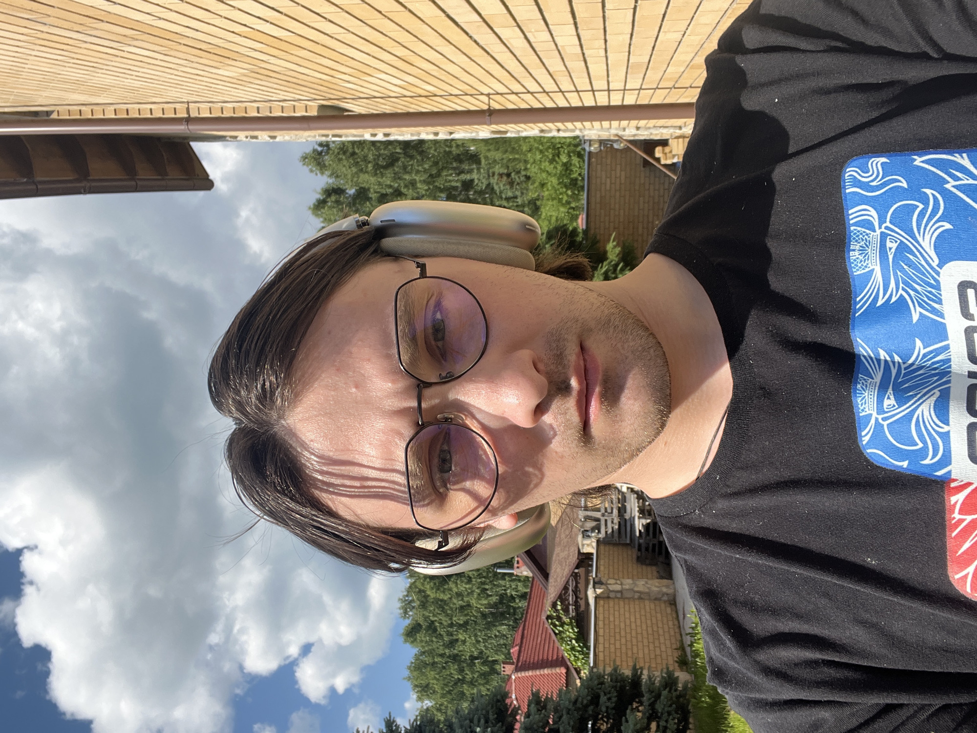 A young man with long dark hair and glasses is taking a selfie outdoors. He is wearing a black t-shirt with a blue and white graphic on the front. He has large silver headphones on. The background shows a brick building on the right, trees, and houses with red roofs in the distance. The sky is partly cloudy. The man appears to be in the city of Orekhovka, Russia, based on the architectural style of the houses and the type of vegetation.