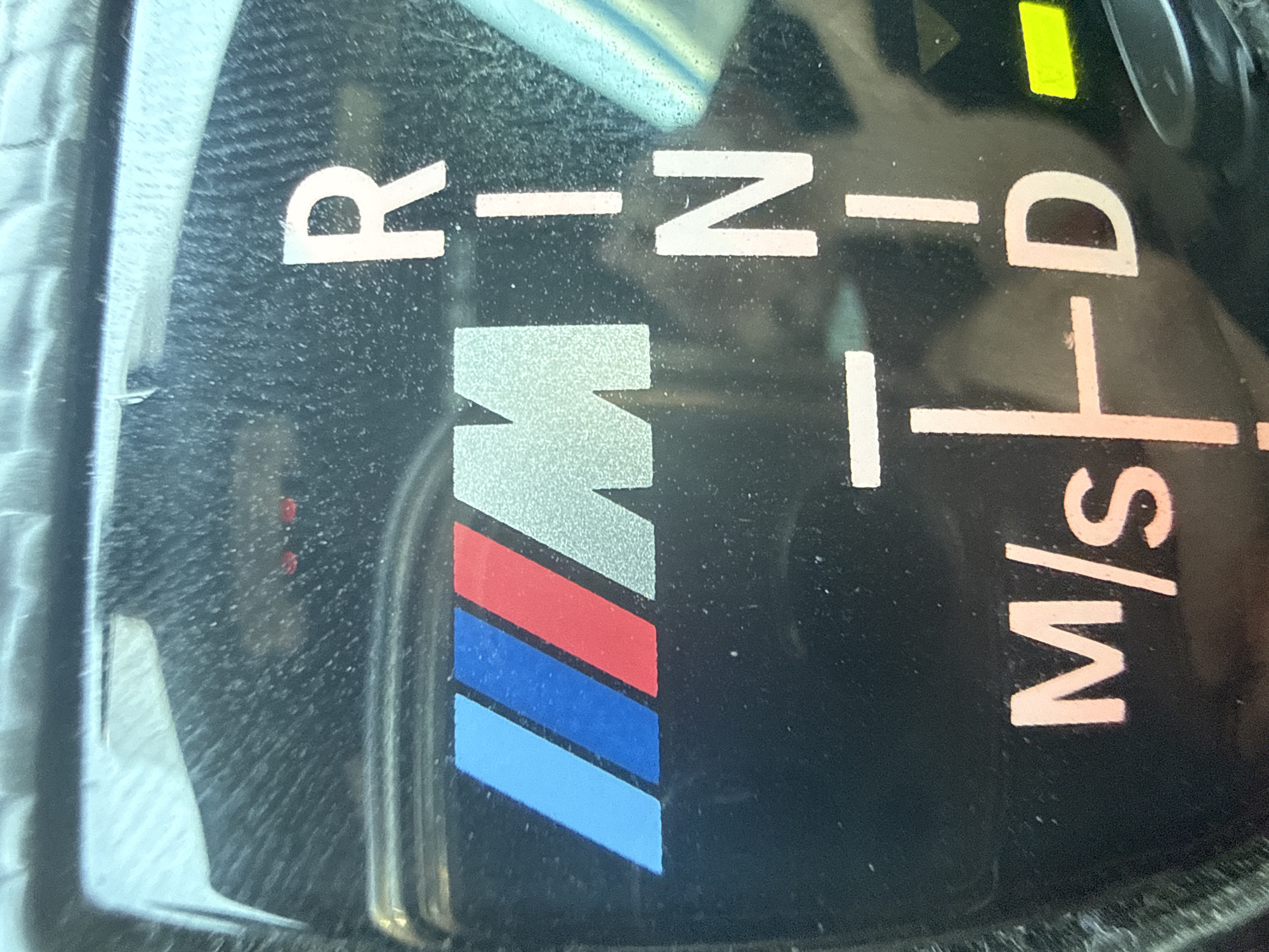 The image displays a close-up of a car's gear selector. The gear indicator shows "R" (Reverse), "N" (Neutral), and "D" (Drive) illuminated. Below "N," there is a selection for "M/S" (Manual/Sport). The BMW M logo is also visible. The environment is the interior of a car, with a black dashboard and some dust particles on the surface. The lighting suggests an indoor setting or a car parked in a shaded area. No people or specific city landmarks are identifiable.
