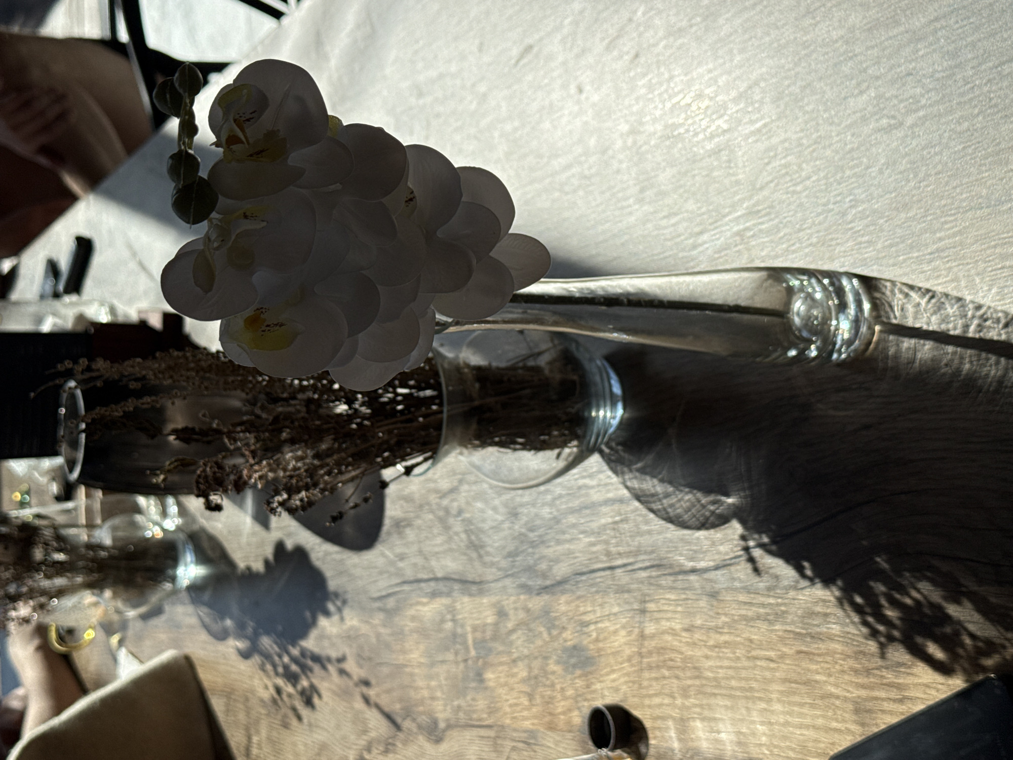 The image displays a static tabletop scene, illuminated by strong, directional sunlight that creates prominent, elongated shadows. In the center, a clear glass bottle or vase lies horizontally, containing white artificial orchids. The orchids are backlit, appearing primarily in silhouette with subtle yellow accents on their petals and dark green buds visible above.
To the left, another clear glass jar, also on its side, holds dried, brown, wispy plants, possibly lavender. Both the orchid vase and the dried plant jar cast distinct shadows across the textured wooden table surface towards the right. The background on the upper left shows a blurred reflection or view, hinting at geometric shapes, possibly from a window or interior structure, and a very indistinct human hand or arm. The upper right background features a bright, subtly textured surface, likely a wall or ceiling, brightly lit.
Small, sparkling bokeh lights are visible around the dried plants. In the bottom right corner, a small, dark cylindrical object and a portion of a dark rectangular item are present. The strong shadows and bright light suggest a sunny day, either in the morning or late afternoon. The overall setting appears to be an indoor space, such as a cafe or home, in Kazan, Russia. No specific activity is depicted, and no visible text is present. The scene conveys a calm and serene atmosphere.