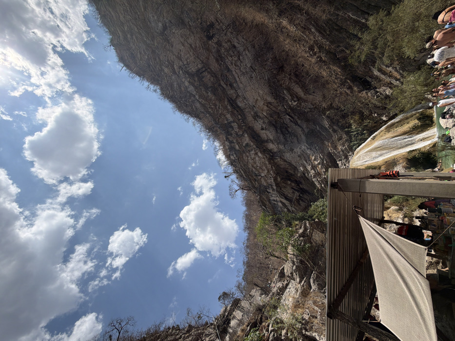 This image depicts a natural canyon environment near San Miguel Atlapulco, Mexico. The upper portion of the frame is dominated by a bright blue sky with scattered white cumulus clouds; the sun is visible in the upper right quadrant, partially obscured by a cloud. Steep, rugged rock faces define the canyon walls, extending from the mid-ground upwards. These cliffs display layered geological formations and are sparsely covered with dry, scrubby vegetation, including bare trees and bushes.
In the mid-ground on the right, a multi-stream waterfall cascades down an inclined, light-colored rock face into turquoise-green pools situated at the base of the canyon. A crowd of approximately 25-35 individuals is dispersed around and within these pools in the lower right of the frame, engaged in recreational water activities, dressed in swimwear or light attire.
In the lower left foreground, a partial man-made structure is visible. It features a corrugated metal roof supported by dark vertical pillars. Beneath this roof, a light-colored canvas tarp is stretched, partially obscuring the interior, where various unspecified items are present. A red and orange life vest hangs from one of the visible pillars.