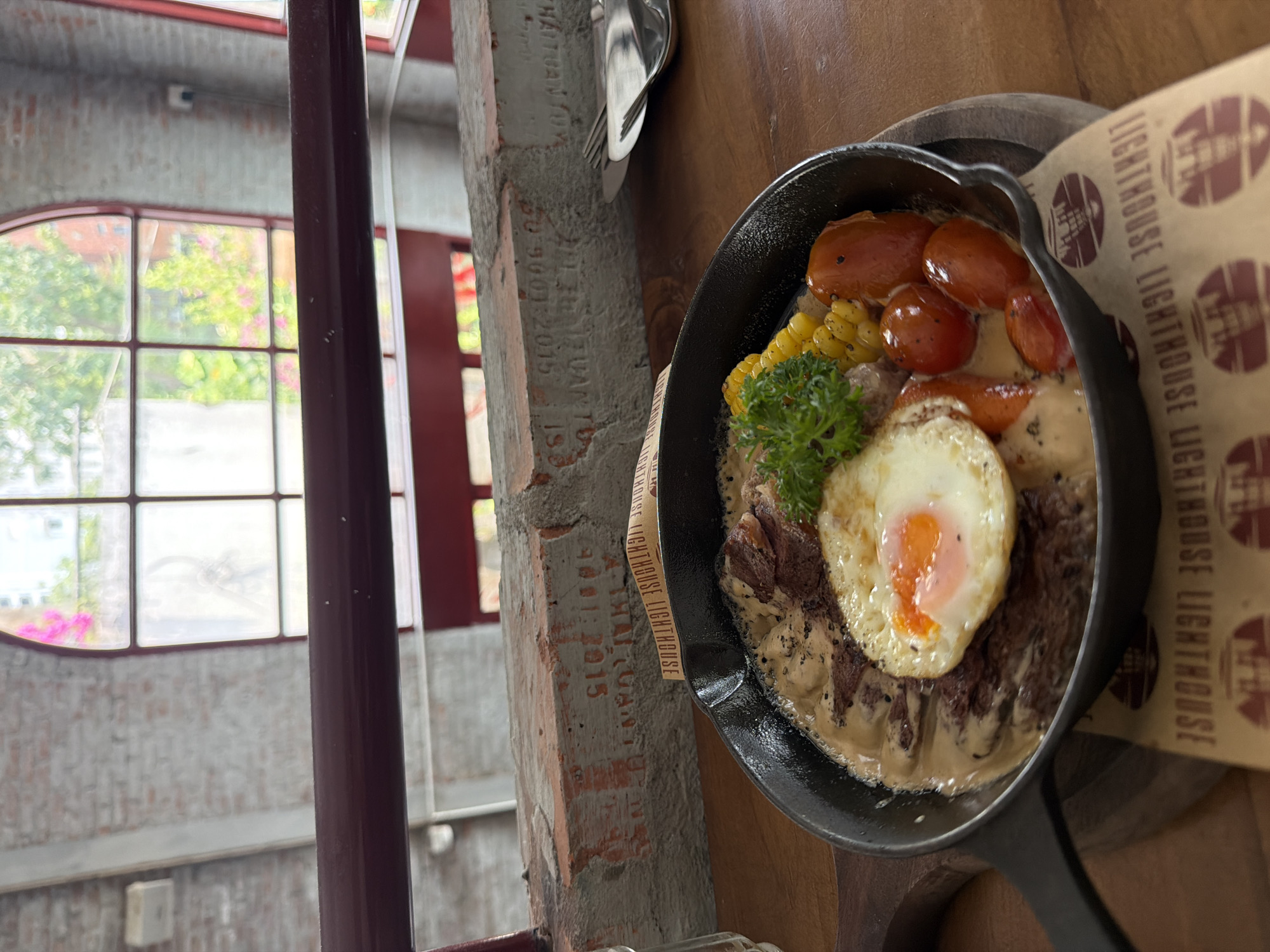 Here is a description of the image:
The image captures a close-up, high-angle view of a cast-iron skillet filled with food, positioned next to a window in what appears to be a restaurant or eatery. The meal consists of a steak topped with a fried egg, garnished with parsley, corn, and several cherry tomatoes. The steak and other ingredients appear to be coated in a creamy sauce. The skillet rests on a wooden board, partly covered by a paper displaying the words "LIGHTHOUSE."
The location seems to be indoors, near a window, allowing the scene to be illuminated by natural light. Outside the window, greenery and a glimpse of the outdoor environment are visible, suggesting a possible urban or suburban setting.
The window has a grid-like pattern and architectural details indicative of a building structure. Next to the window, there's a brick column or wall section with printed words "HAITUAN TUDANT9001:2015 50-9001:2015."
Overall, the scene seems to portray a culinary moment in a restaurant setting, likely in Da Nang, Vietnam, as indicated by the location "Lighthouse." The image conveys the meal and a partial view of the environment, creating a sense of dining ambiance.