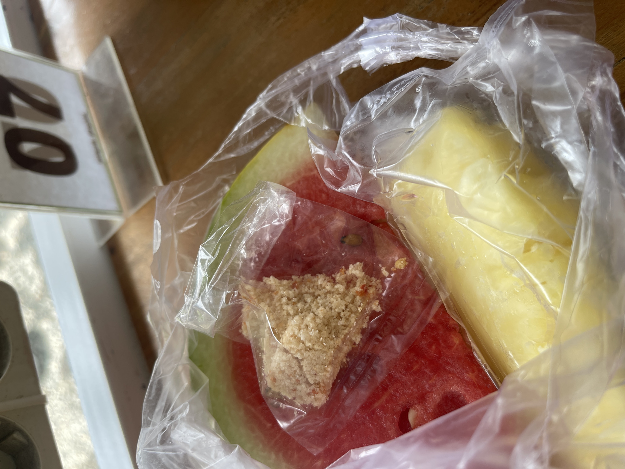 A close-up shot captures a plastic bag containing pieces of fruit, likely watermelon and pineapple, placed on a wooden table. A small portion of a numbered table marker, displaying "07", is visible in the background, suggesting a restaurant or dining establishment setting. The lighting indicates an indoor environment, possibly with natural light filtering in. The scene appears to be within the city of Pulokiton, Indonesia.
