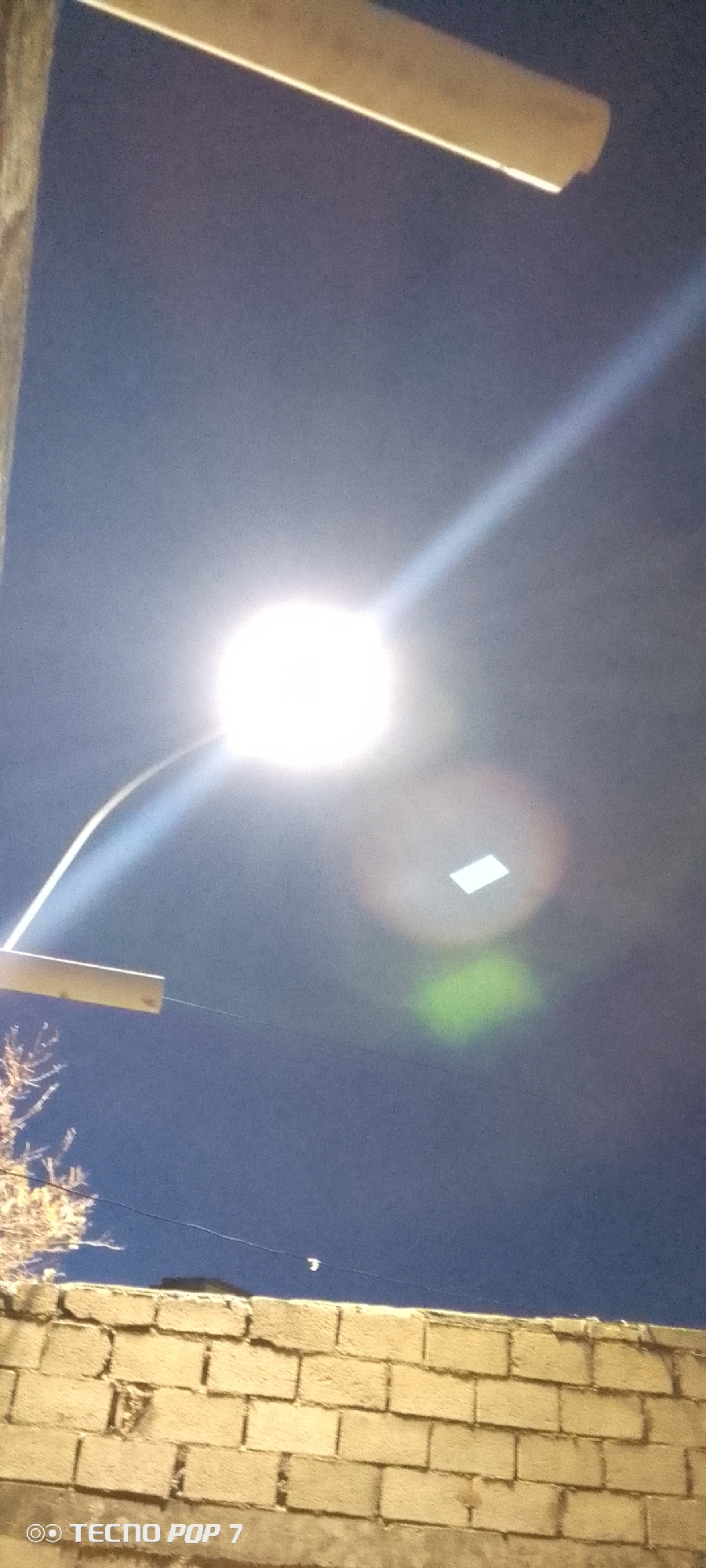 In Altagracia, Venezuela, at night, a bright streetlamp illuminates a dark sky. Below the lamp, a curved metal arm supports the light fixture, which is angled upwards. A horizontal wooden beam is visible in the upper left portion of the frame, casting a shadow. The sky is a deep blue, with a faint white streak emanating from the lamp. In the middle of the sky, there is a faint, rectangular-shaped reflection. To the right of the reflection, a green glow is visible. In the lower portion of the image, a brick wall runs horizontally across the frame, with some sparse branches of a tree visible above it on the left. A small, white object, possibly a distant bird or drone, is visible in the sky above the wall. The bottom of the image displays a watermark reading "TECNO POP 7".
