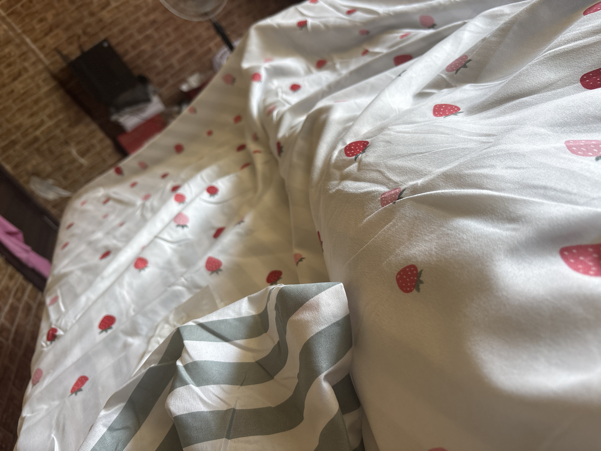 A close-up shot of a white duvet cover adorned with a pattern of red strawberries. The duvet cover is rumpled, suggesting it has been recently used. To the left, a pillow with green and white stripes is partially visible, creating a contrast with the strawberry pattern. The background is blurred, but appears to be a brick wall and some storage containers, suggesting an indoor setting. The lighting is natural and appears to be coming from the right side of the image.